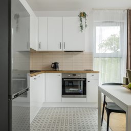 GB Foods Grzegorz Bandzarewicz - Jasna kuchnia z białymi szafkami, drewnianym blatem i wzorzystą podłogą. Na stole misa z owocami, w tle okno z zasłonami i fragment salonu.