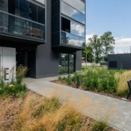 Nowoczesny budynek apartamentowy z balkonami, zagospodarowanym terenem zielonym i ławką. Minimalistyczna architektura,  numer '1' przy wejściu.