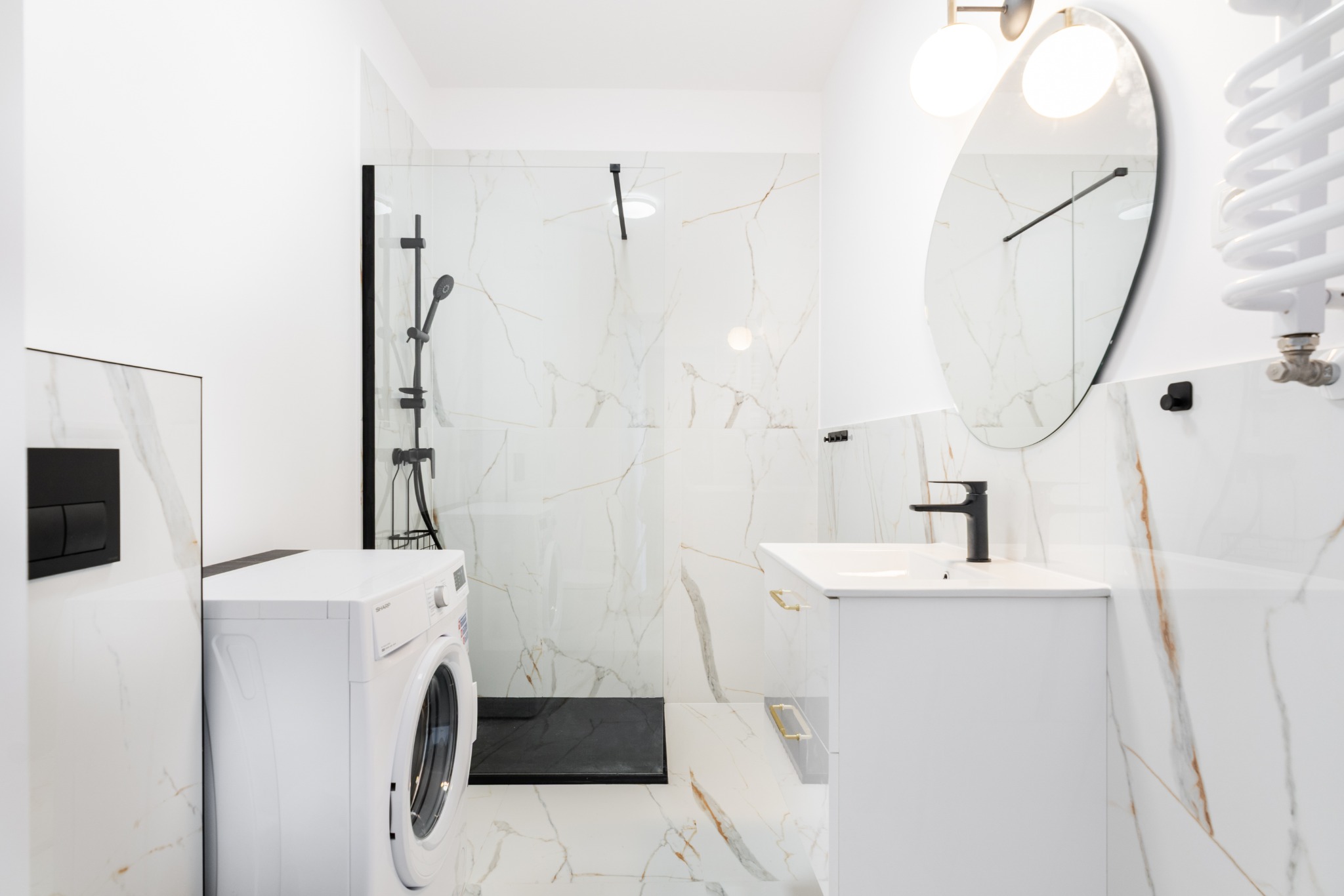 Nowoczesna łazienka w bieli z marmurowym wzorem, czarną armaturą i kabiną prysznicową typu walk-in. Widoczna pralka i owalne lustro z oświetleniem.