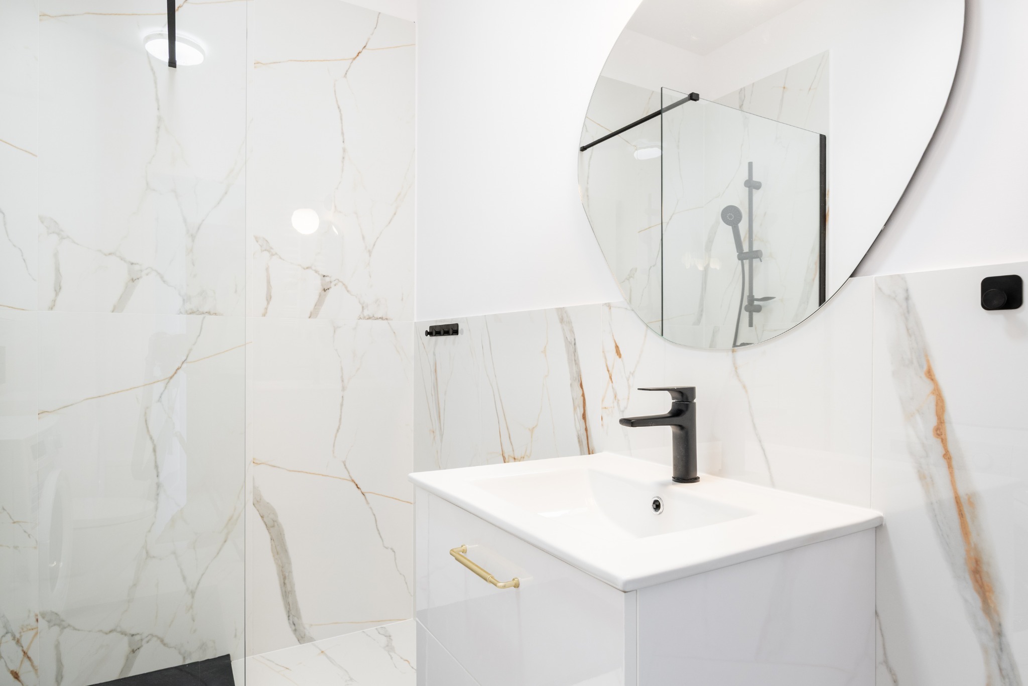 Nowoczesna łazienka z białymi płytkami imitującymi marmur, czarną armaturą i okrągłym lustrem. Złote uchwyty szafki dodają elegancji.