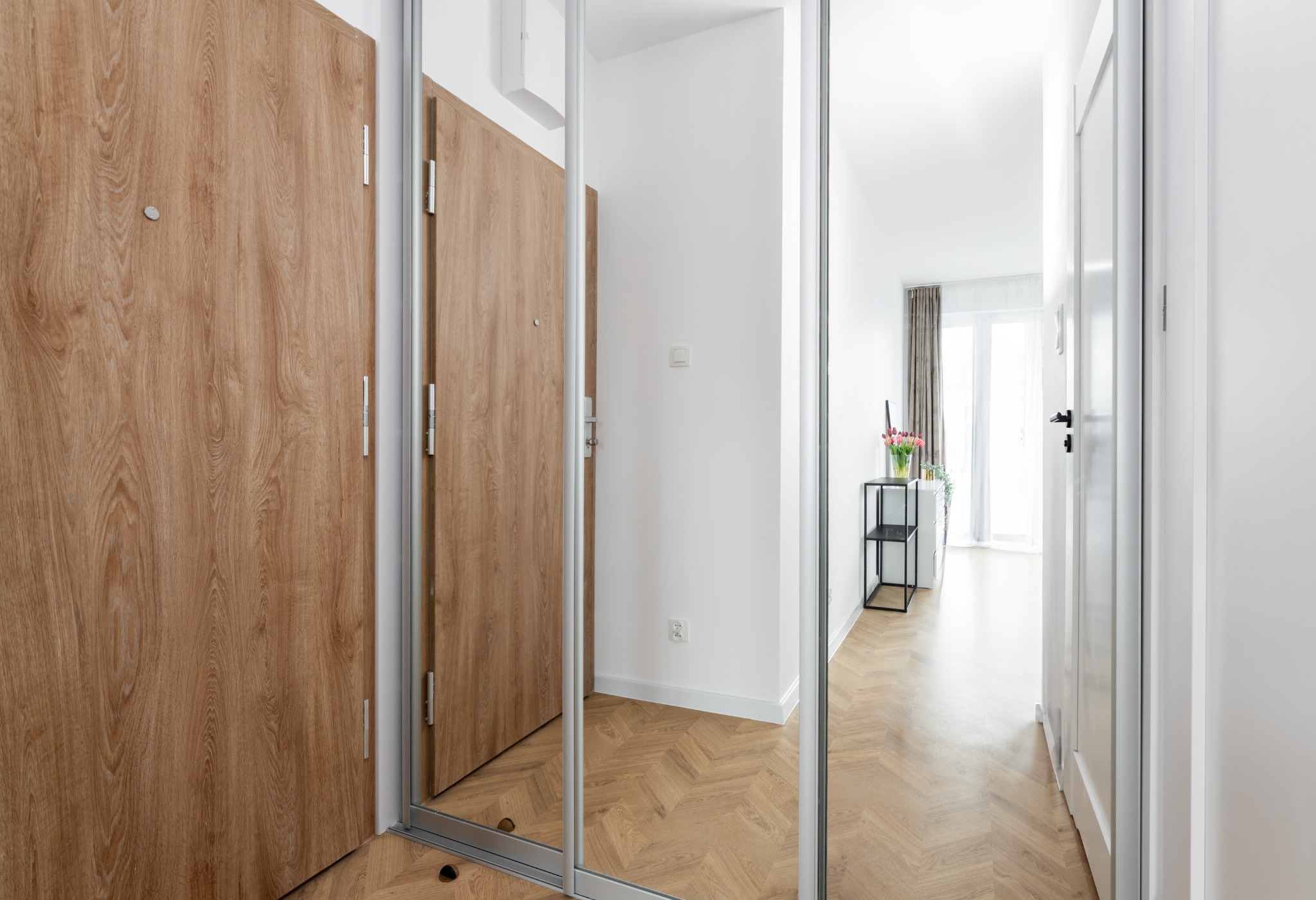 Nowoczesne wnętrze mieszkania z drewnianą podłogą w jodełkę, lustrzanymi drzwiami szafy i widokiem na jasny pokój z kwiatami w wazonie.