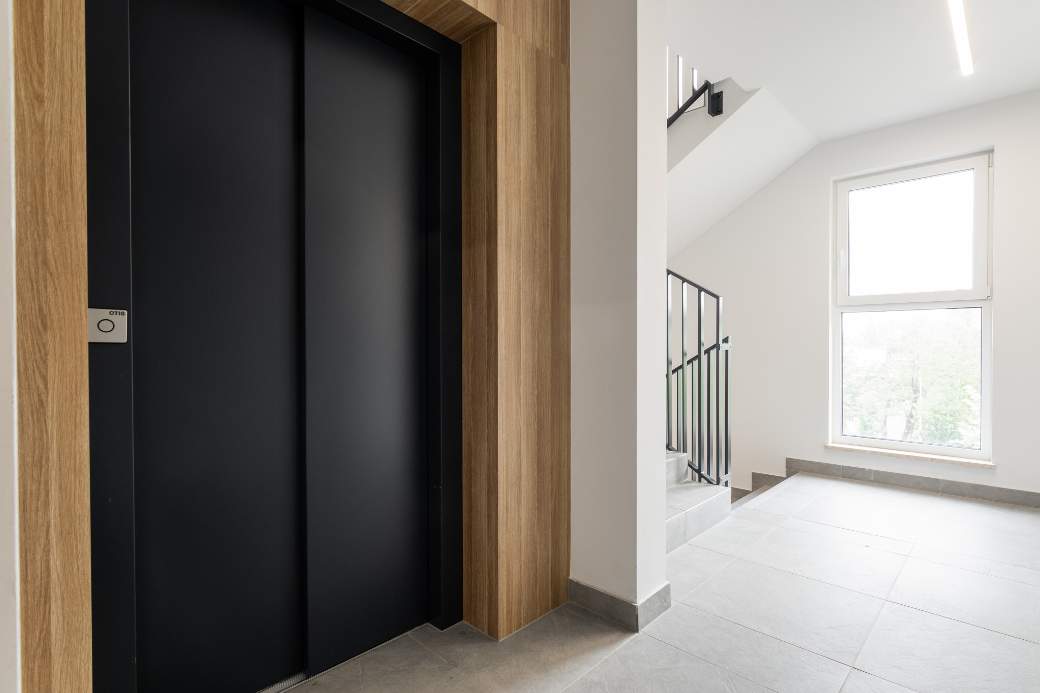 Nowoczesny hol wejściowy: czarna winda Otis, drewniane wykończenie i jasne płytki podłogowe. Minimalistyczne schody i okno w tle. Oświetlenie LED.