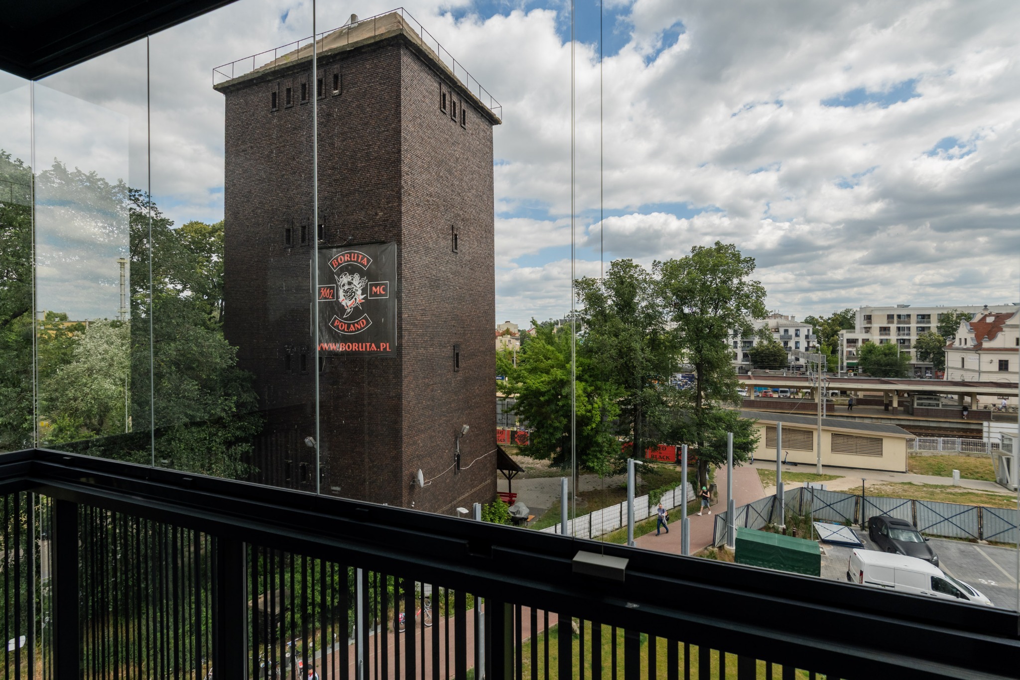 Widok z balkonu na ceglaną wieżę z logiem Boruta MC, drzewa, budynki i parking w tle. Ujęcie przez szklaną balustradę balkonu.