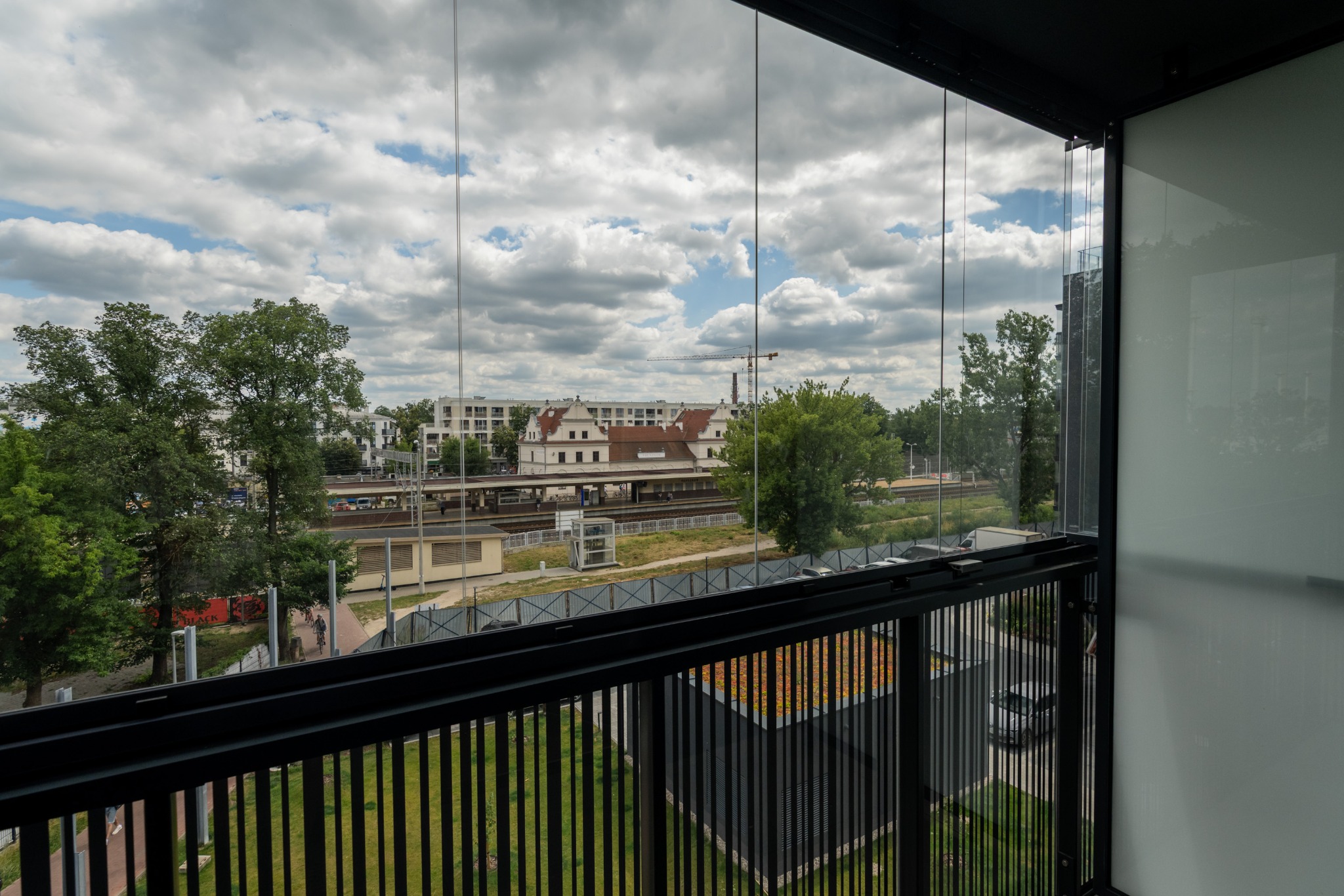 Zabudowany balkon z widokiem na miasto i stację kolejową w słoneczny dzień. Nowoczesne przeszklenia i czarne barierki. Widok z góry.