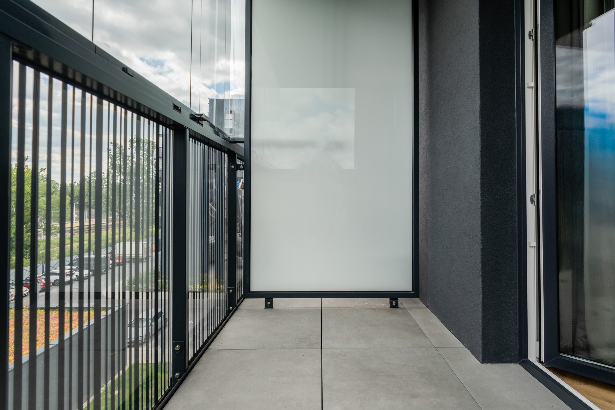 Nowoczesny balkon z szarymi płytkami, czarną balustradą z pionowymi szczebelkami i szklaną osłoną, z widokiem na zieleń i budynki w tle.