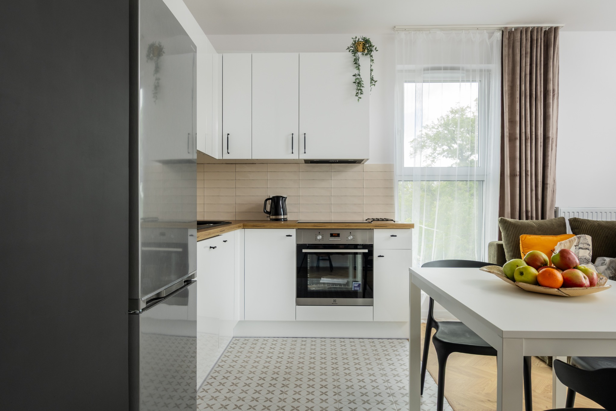 Jasna kuchnia z białymi szafkami, drewnianym blatem i wzorzystą podłogą. Na stole misa z owocami, w tle okno z zasłonami i fragment salonu.