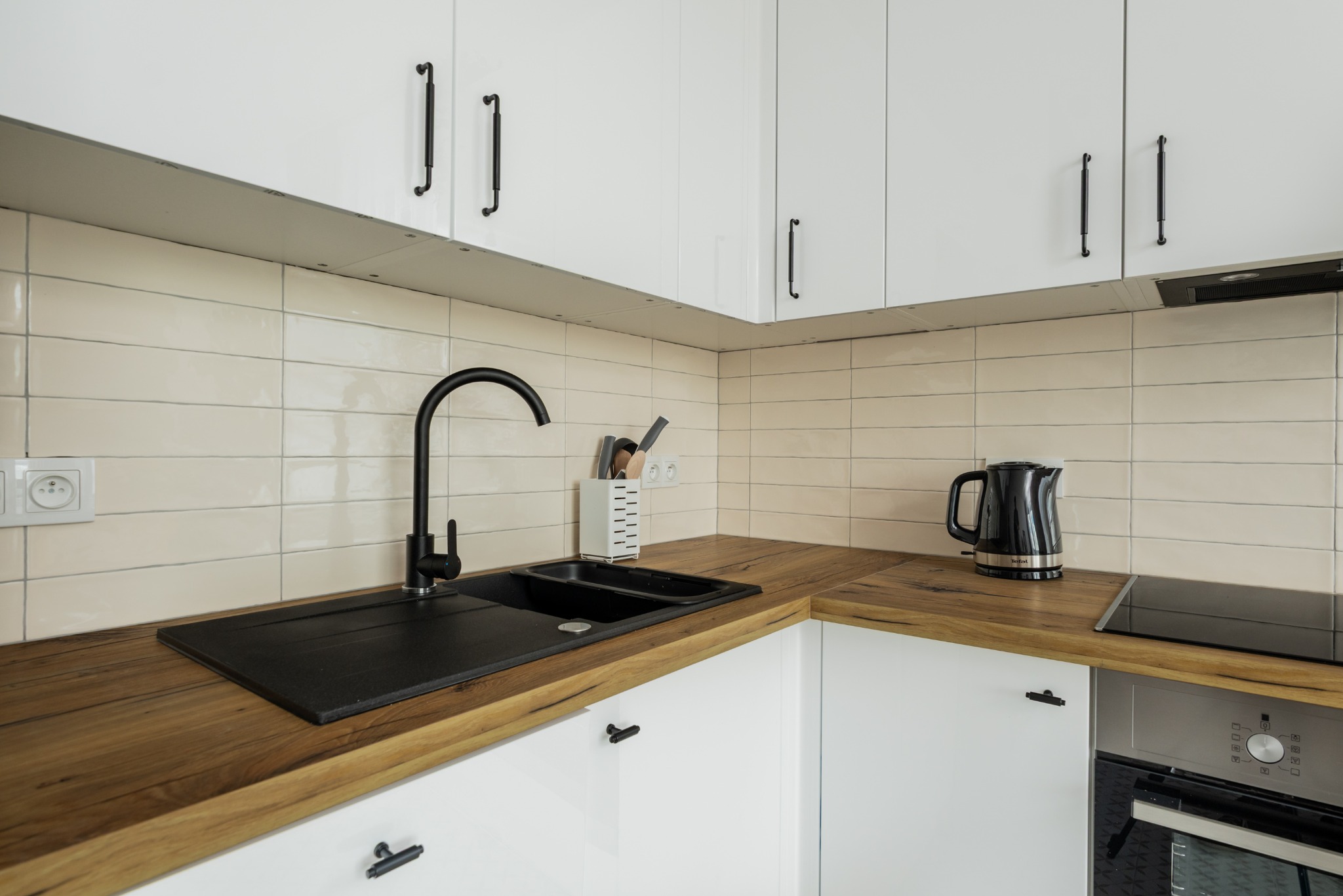 Nowoczesna kuchnia z drewnianym blatem, czarnym zlewem i armaturą. Białe szafki z podłużnymi uchwytami. Na blacie czajnik i płyta indukcyjna. Beżowe płytki na ścianie.