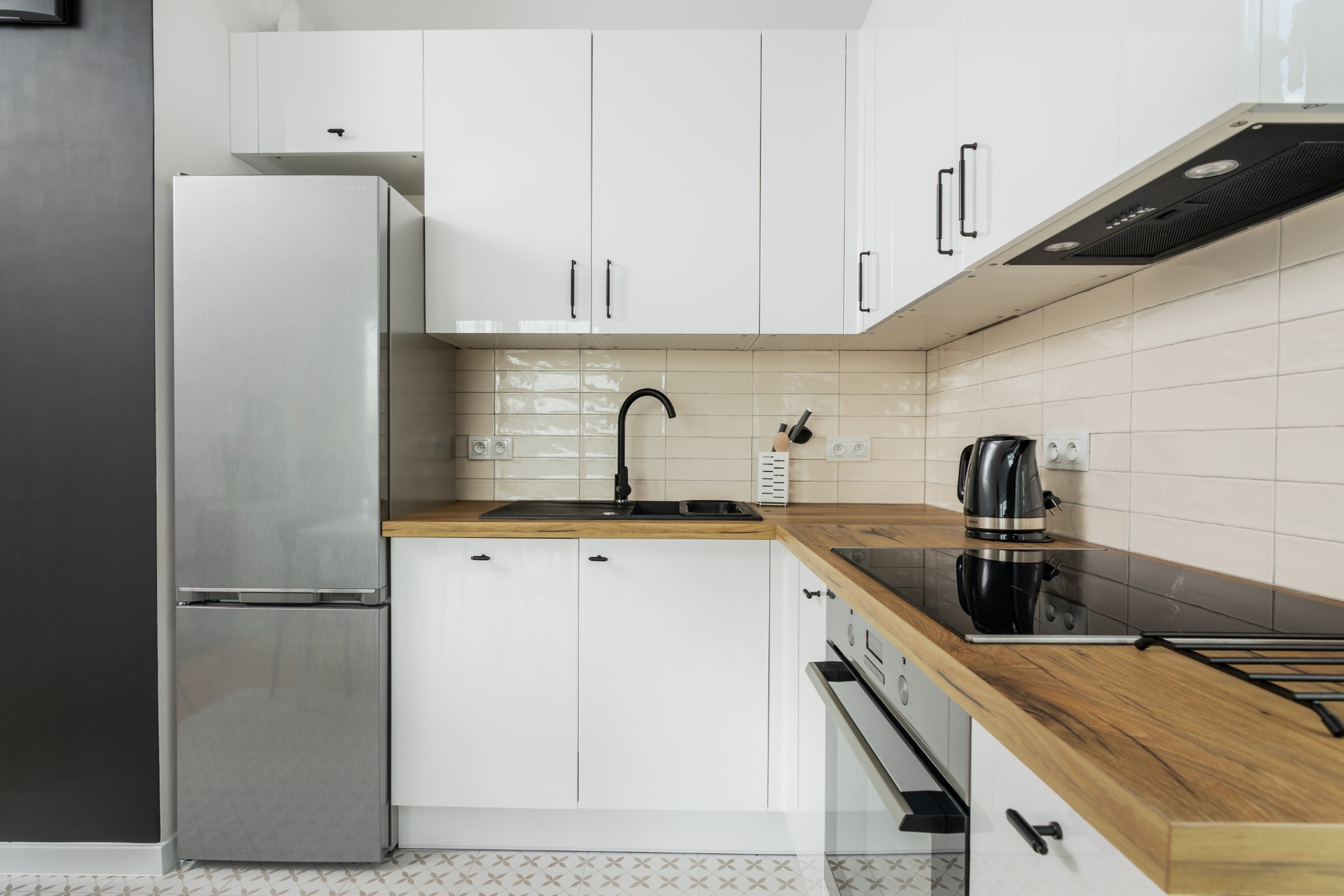 Jasna kuchnia z drewnianym blatem i białymi szafkami. Widoczny srebrny lodówka, czarny zlew i czajnik. Podłoga z wzorem w kwiaty. Nowoczesny design.