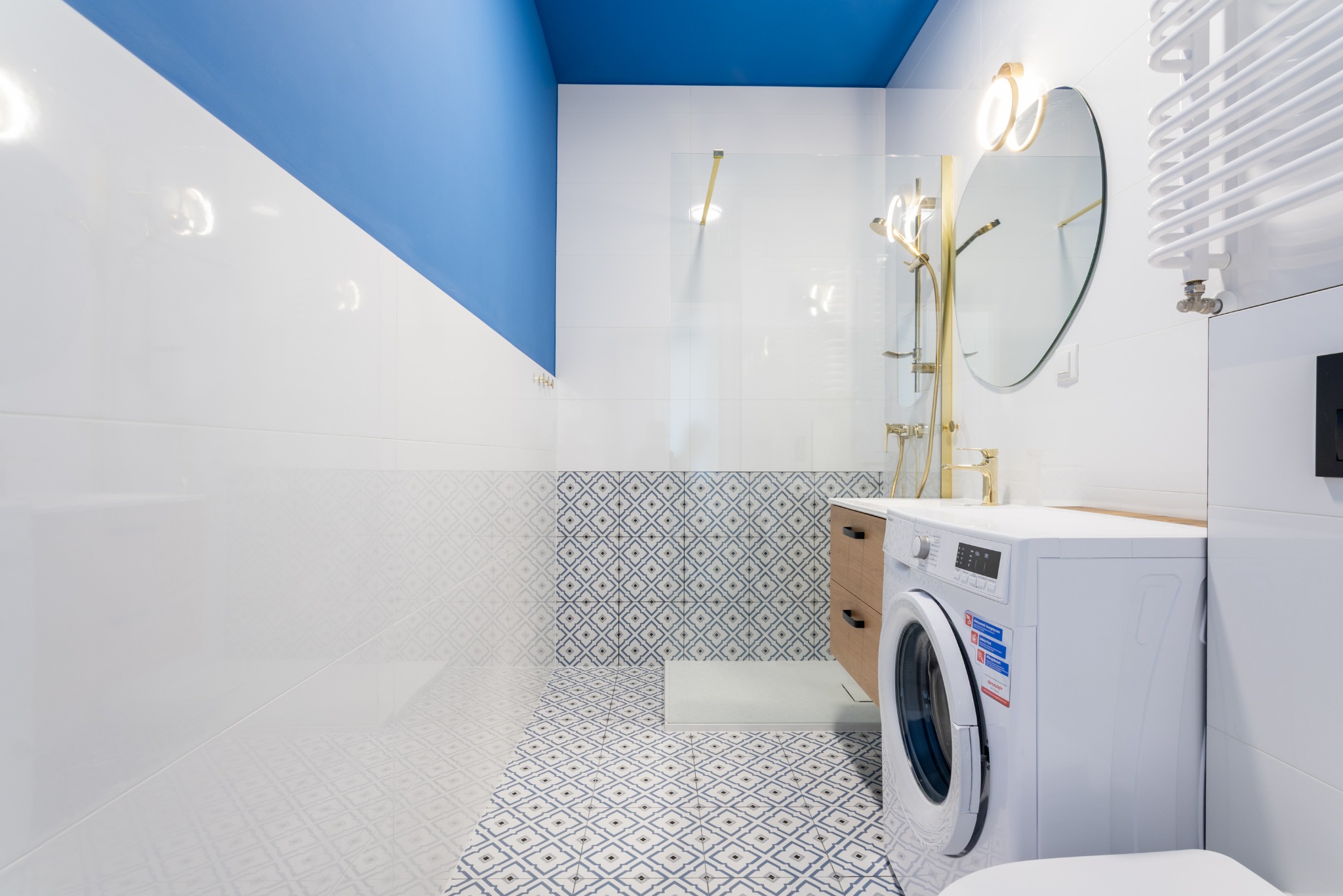 Nowoczesna łazienka z niebieskim sufitem, białymi płytkami i wzorzystą podłogą. Złota armatura, okrągłe lustro z podświetleniem i pralka w minimalistycznym stylu.
