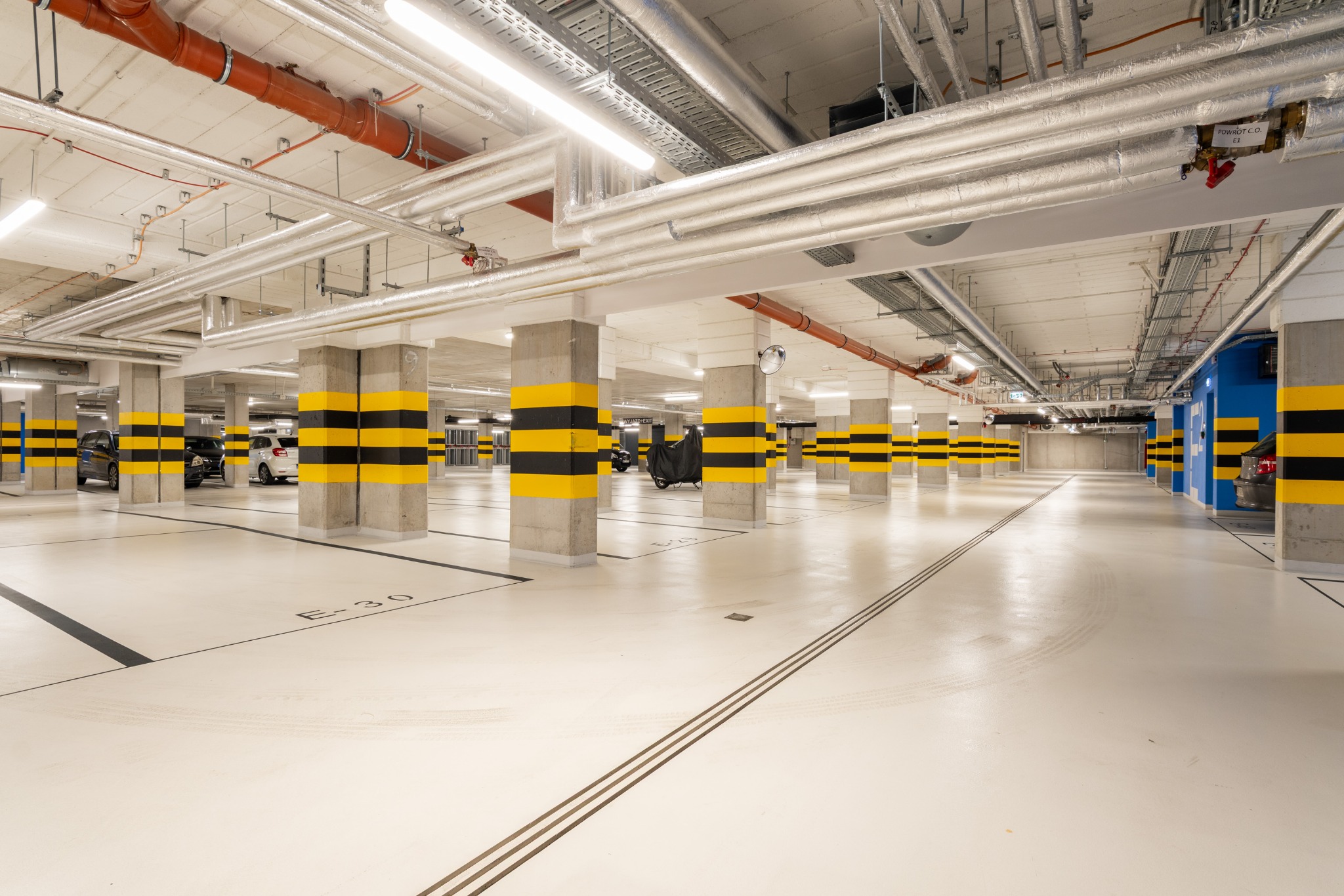 Nowoczesny garaż podziemny z oznaczonymi miejscami parkingowymi, kolumny w pasy żółto-czarne, sufit z instalacjami. Czysta, jasna przestrzeń.