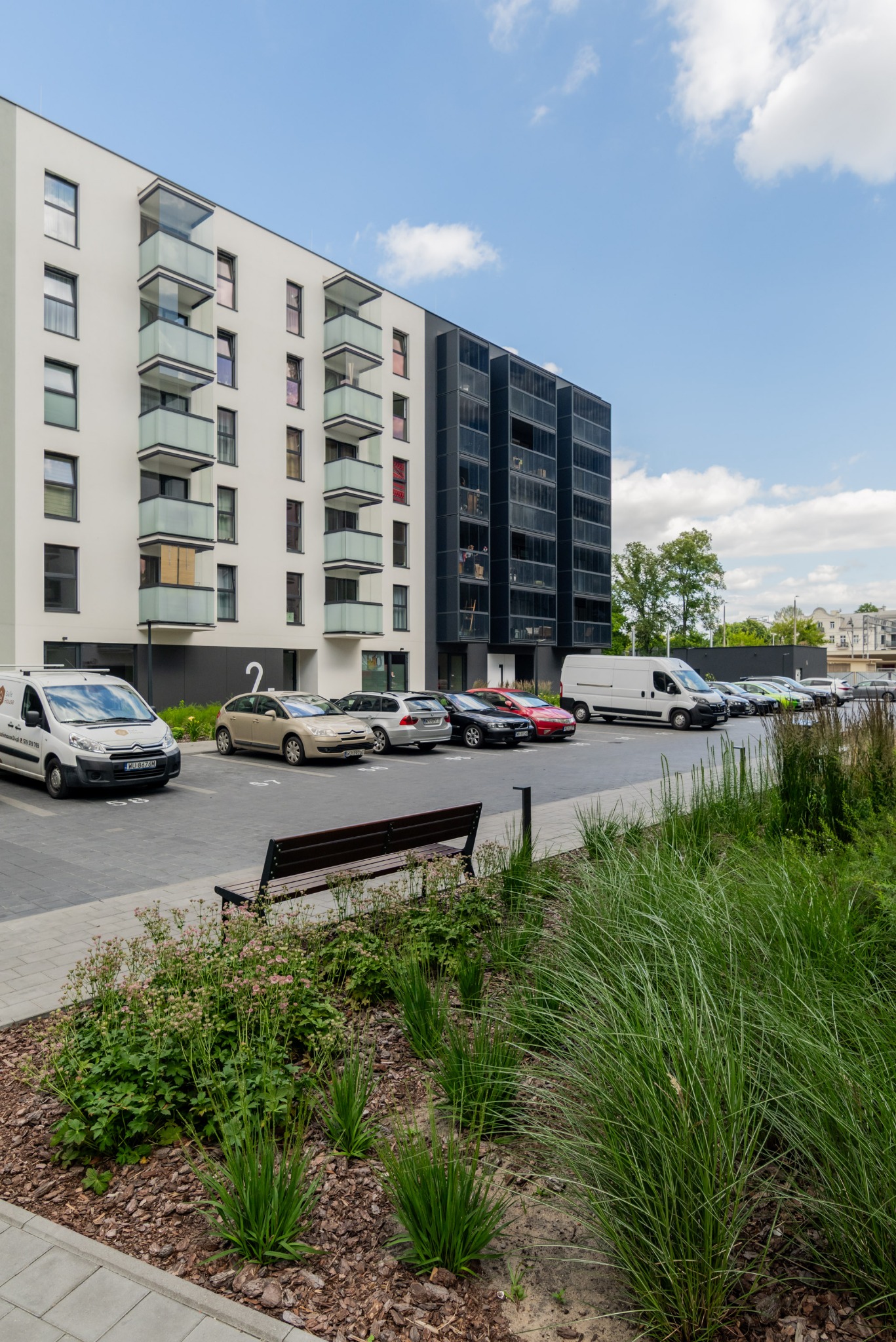 Nowoczesny budynek apartamentowy z balkonami i miejscami parkingowymi, otoczony zielenią. Widoczny fragment chodnika i ławka. Słoneczny dzień.