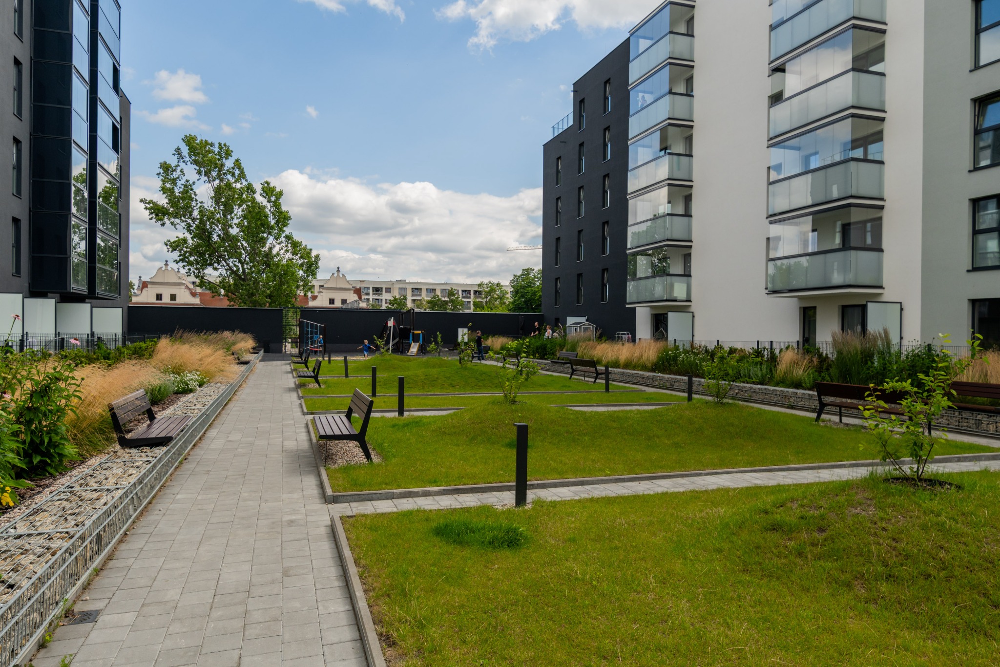Osiedle z nowoczesną architekturą: bloki z balkonami, zielony dziedziniec z ławkami i placem zabaw w tle, błękitne niebo z chmurami. Letni dzień.