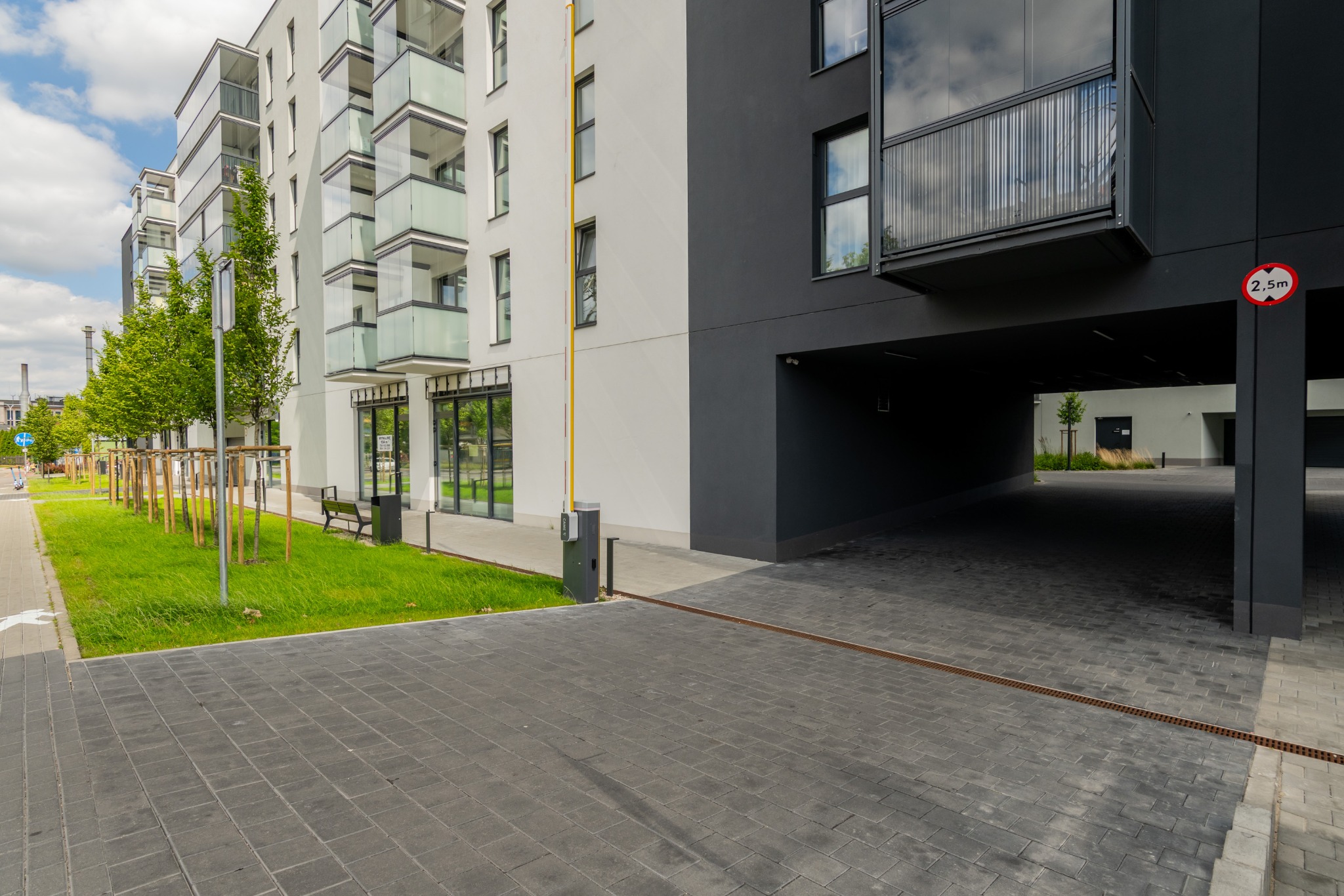 Nowoczesny budynek apartamentowy w Dokudowie Drugim z przeszklonymi balkonami i wjazdem do garażu, widok z poziomu chodnika z kostki brukowej.