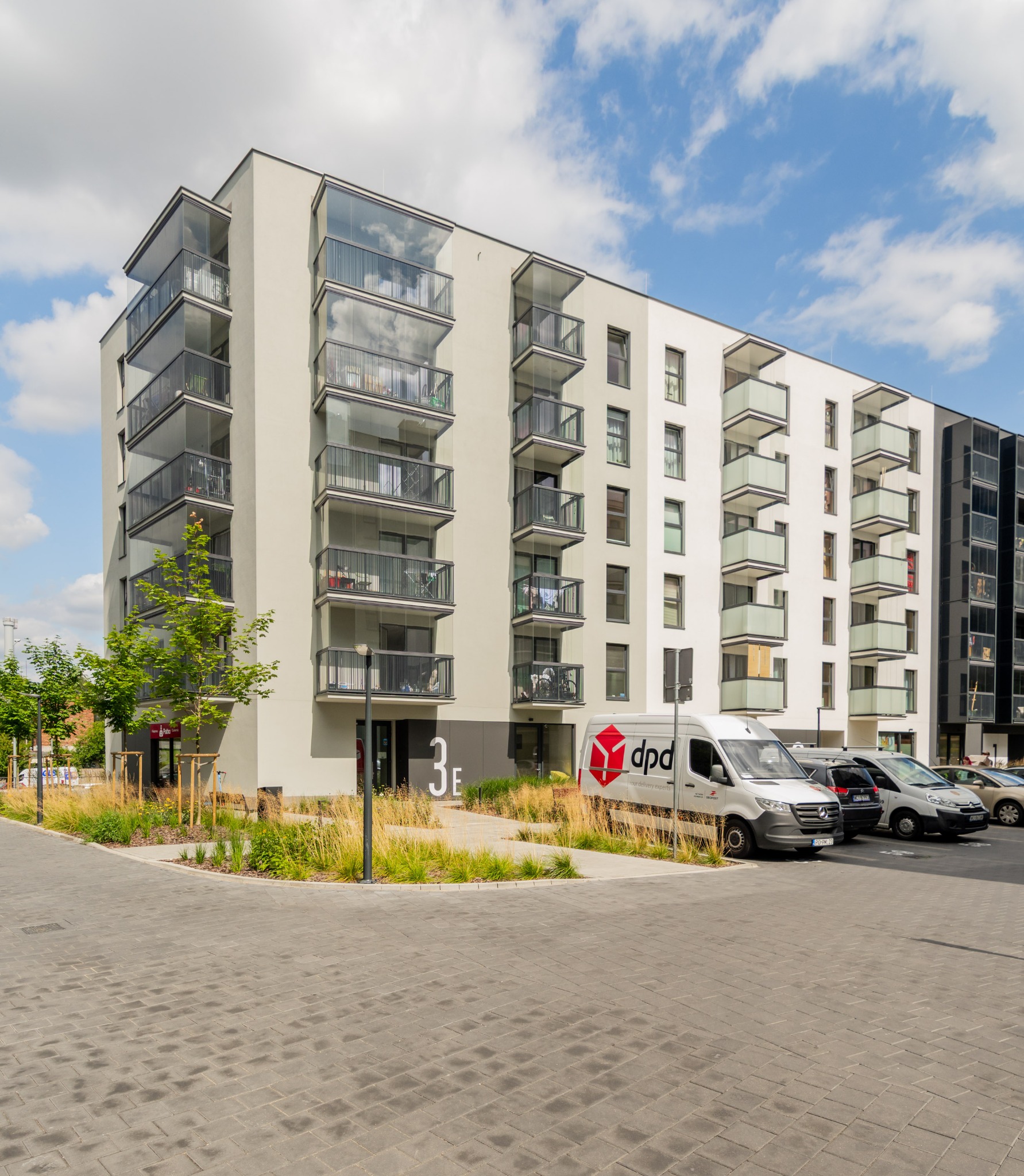 Nowoczesny budynek apartamentowy z balkonami i przeszklonymi loggiami, na parkingu zaparkowane samochody, w tym van z logo firmy kurierskiej.