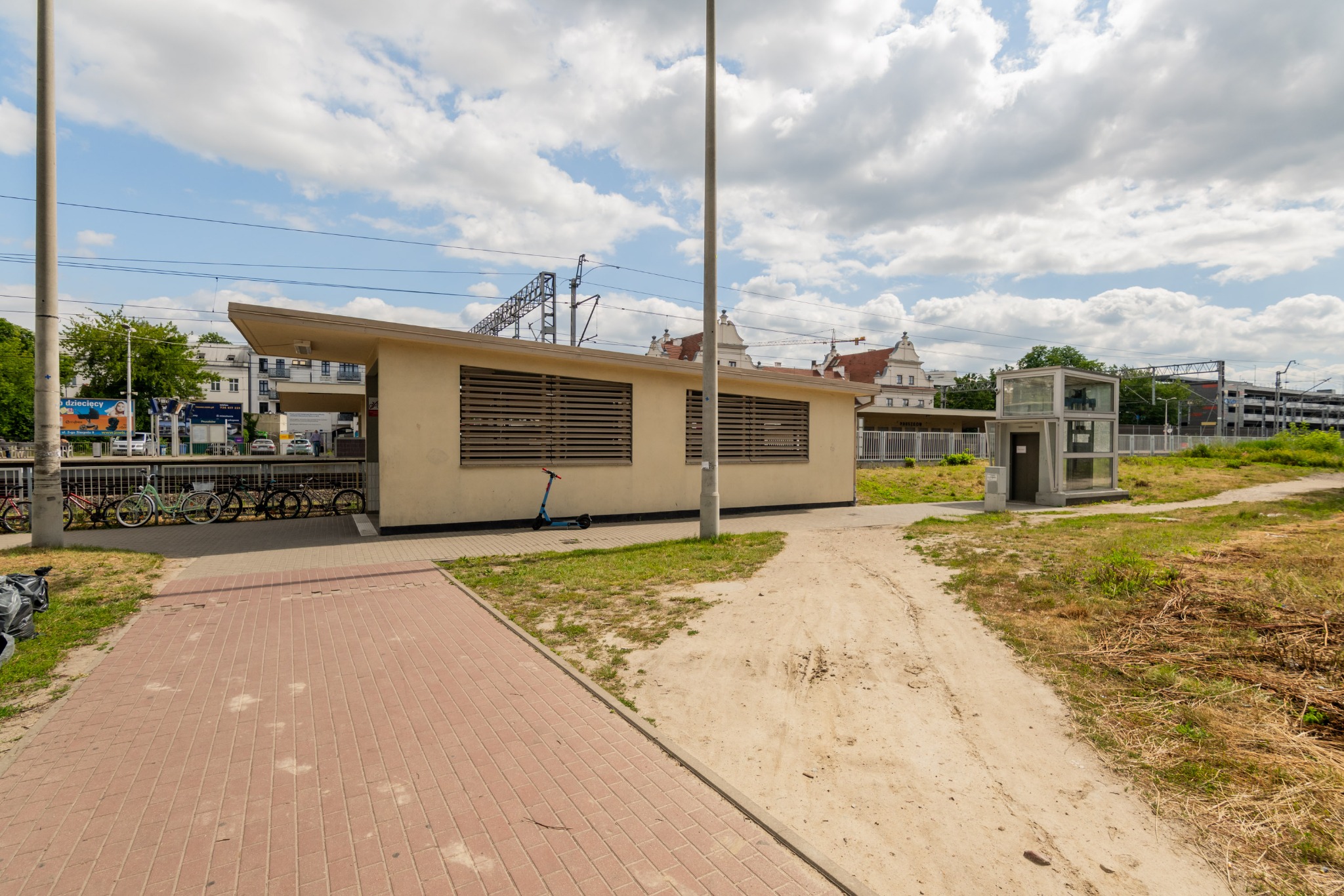 Budynek stacji kolejowej z rowerami i windą dla niepełnosprawnych w słoneczny dzień. Widok na peron i okoliczne budynki. Na pierwszym planie chodnik z kostki.