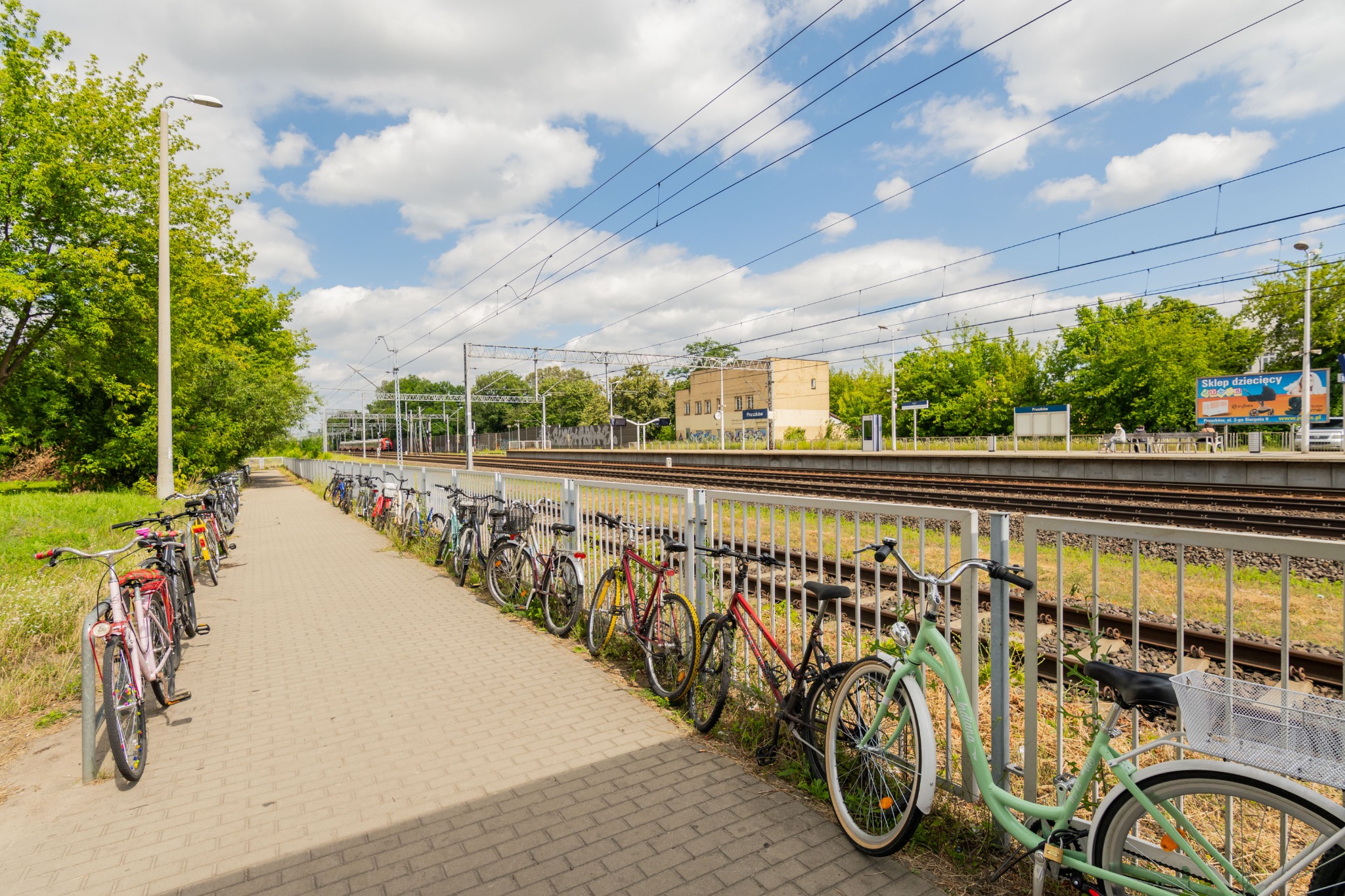 Rząd rowerów przypiętych do barierek wzdłuż chodnika przy stacji kolejowej w słoneczny dzień. Widoczne tory, budynek stacji i zieleń.