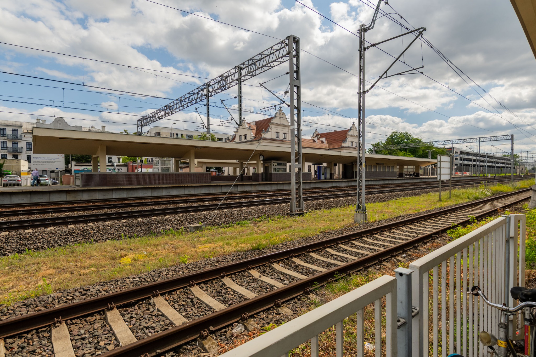 Peron kolejowy z budynkiem dworca w tle, widok zza metalowego ogrodzenia. Tory kolejowe z zielenią, słupy trakcyjne i pochmurne niebo.