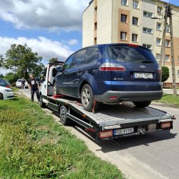 SZYM CARS SZYMON GŁUCHOWSKI - Laweta z niebieskim Fordem S-Max na platformie, zaparkowana na poboczu ulicy w słoneczny dzień. W tle blok mieszkalny i osoba w czarnej koszulce.