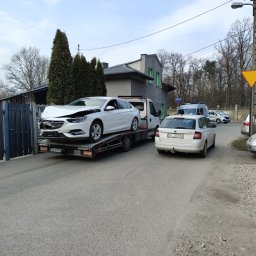 SZYM CARS SZYMON GŁUCHOWSKI - Biała laweta z uszkodzonym białym Oplem Insignia na platformie, zaparkowana na ulicy obok zaparkowanych aut, w tle budynek i drzewa. Widoczny znak drogowy.