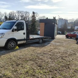 SZYM CARS SZYMON GŁUCHOWSKI - Biała laweta Iveco z żółtym klinem pod kołem przewozi czarny pawilon z elementami drewnianymi, na tle drzew i budynków w słoneczny dzień.