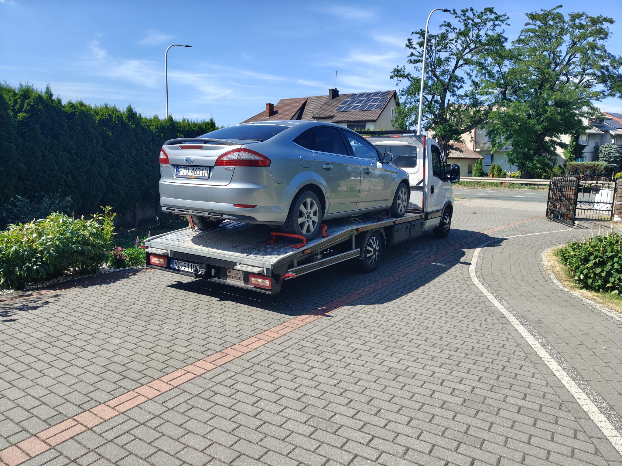 Srebrny Ford Mondeo na platformie lawety, zaparkowanej na brukowanym podjeździe przed domem. Widoczny tył samochodu i fragment białej kabiny lawety.