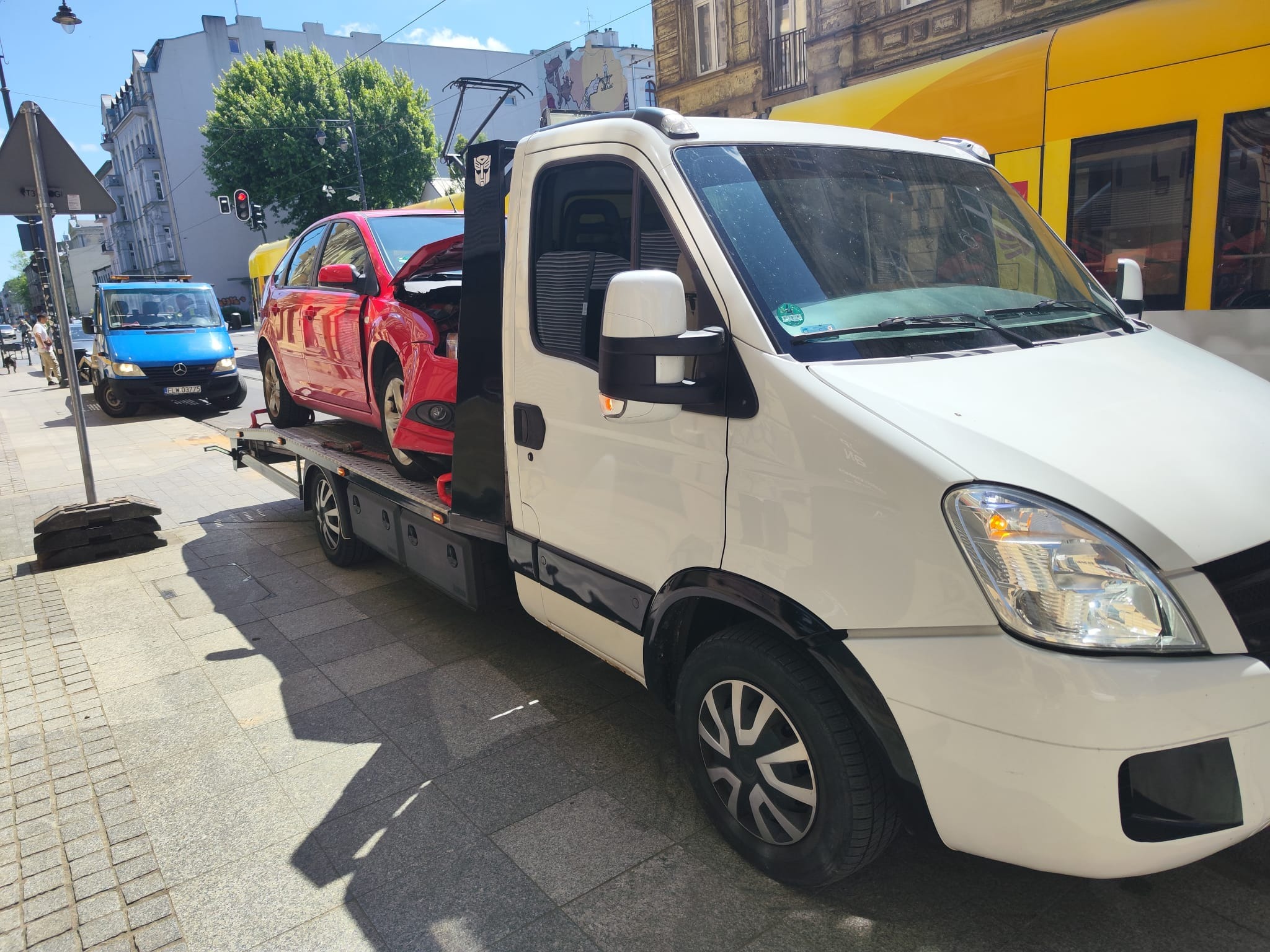 Biała laweta z czerwonym samochodem na platformie, zaparkowana na chodniku w słoneczny dzień, z tramwajem w tle. Na drzwiach lawety naklejka z logiem Transformers.