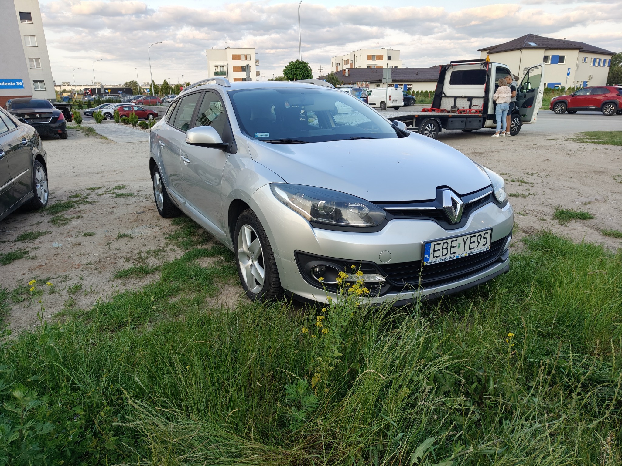 Srebrny Renault Megane kombi na trawie, z widoczną lawetą w tle. Tablica rejestracyjna EBE YE95. Ujęcie z dołu, podkreślające wysokość trawy.