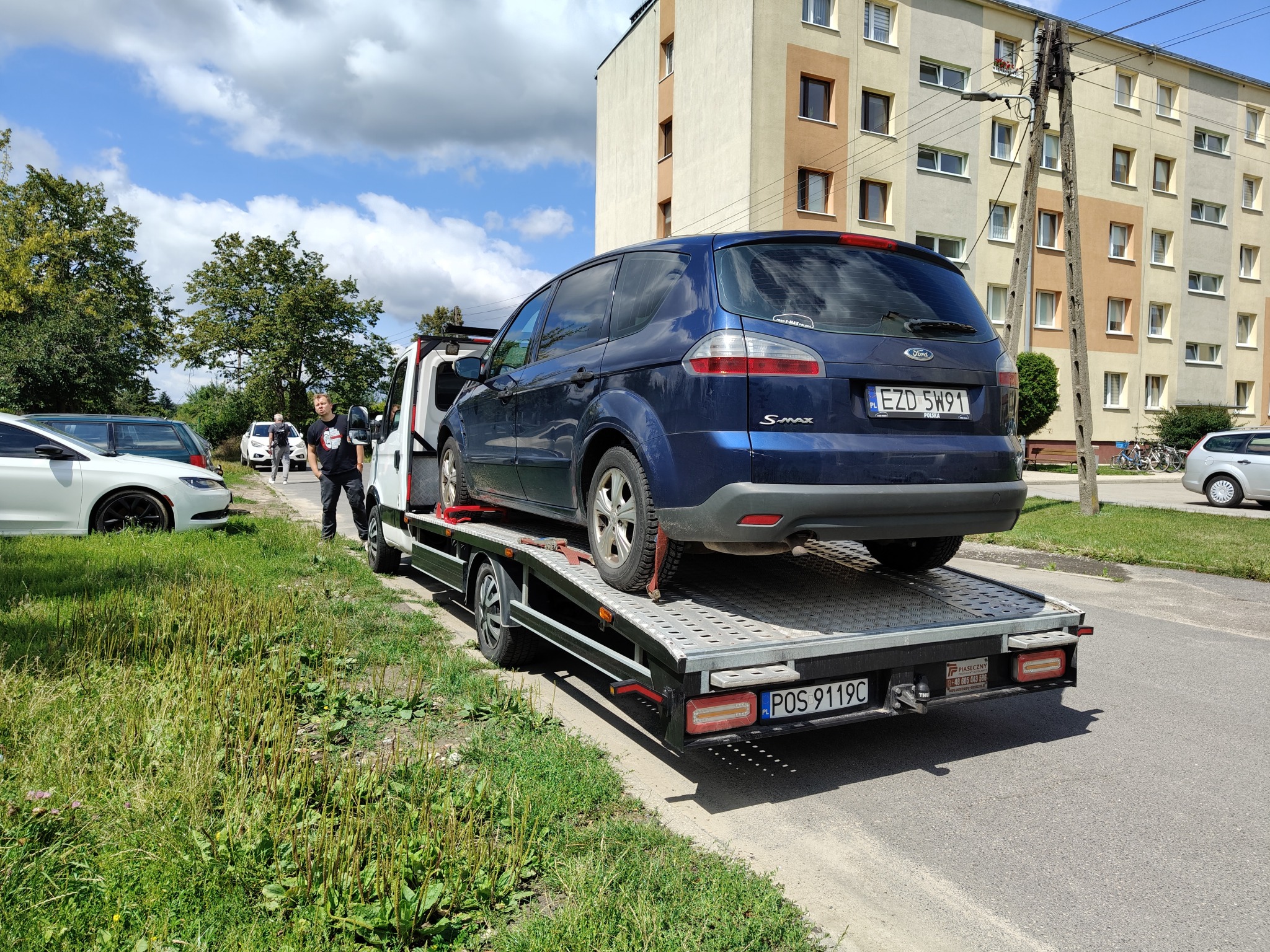 Laweta z niebieskim Fordem S-Max na platformie, zaparkowana na poboczu ulicy w słoneczny dzień. W tle blok mieszkalny i osoba w czarnej koszulce.