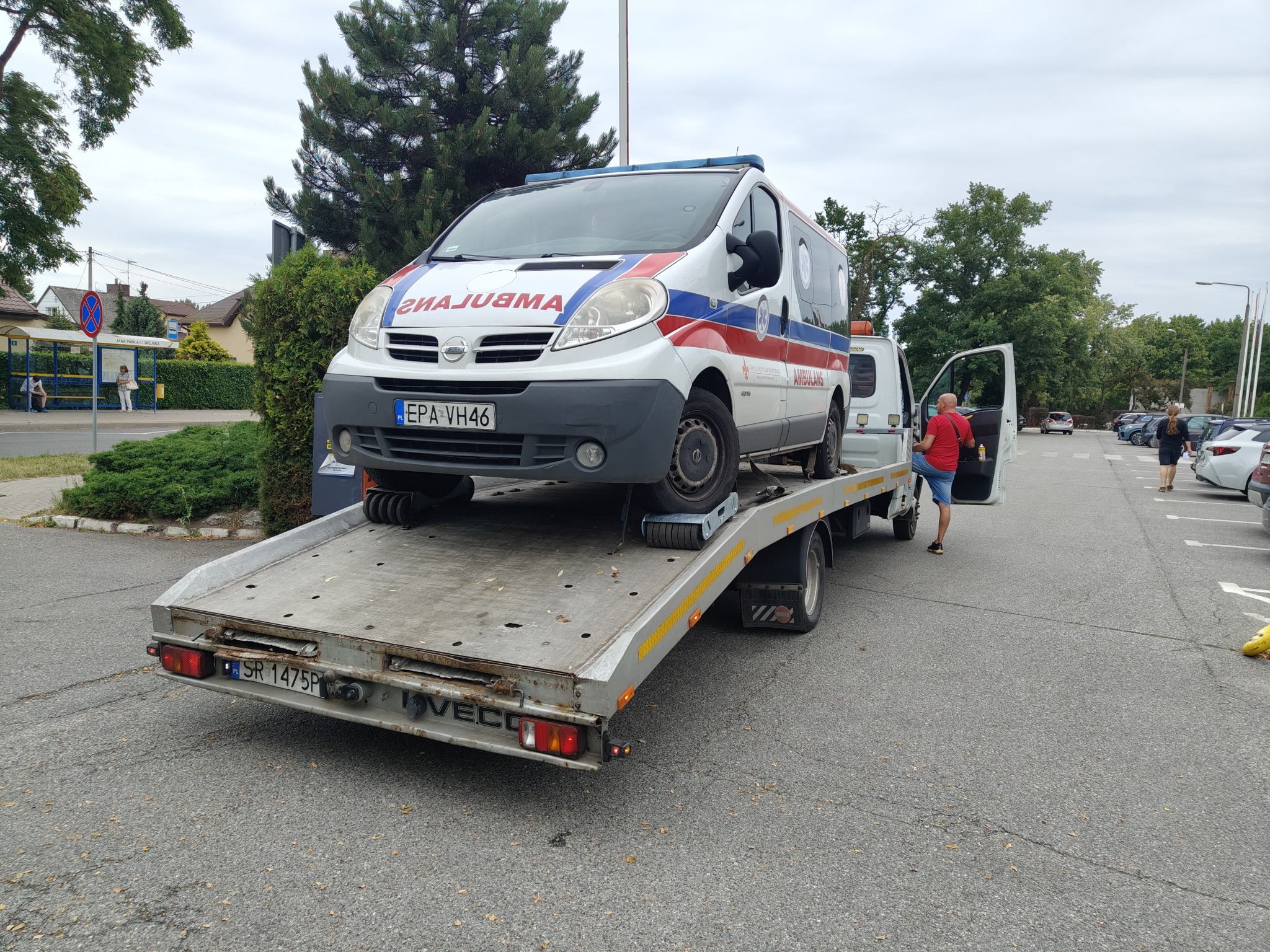Laweta z ambulansem na platformie, otwarte drzwi kabiny, mężczyzna w czerwonej koszulce. Parking z innymi pojazdami w tle, dzień.