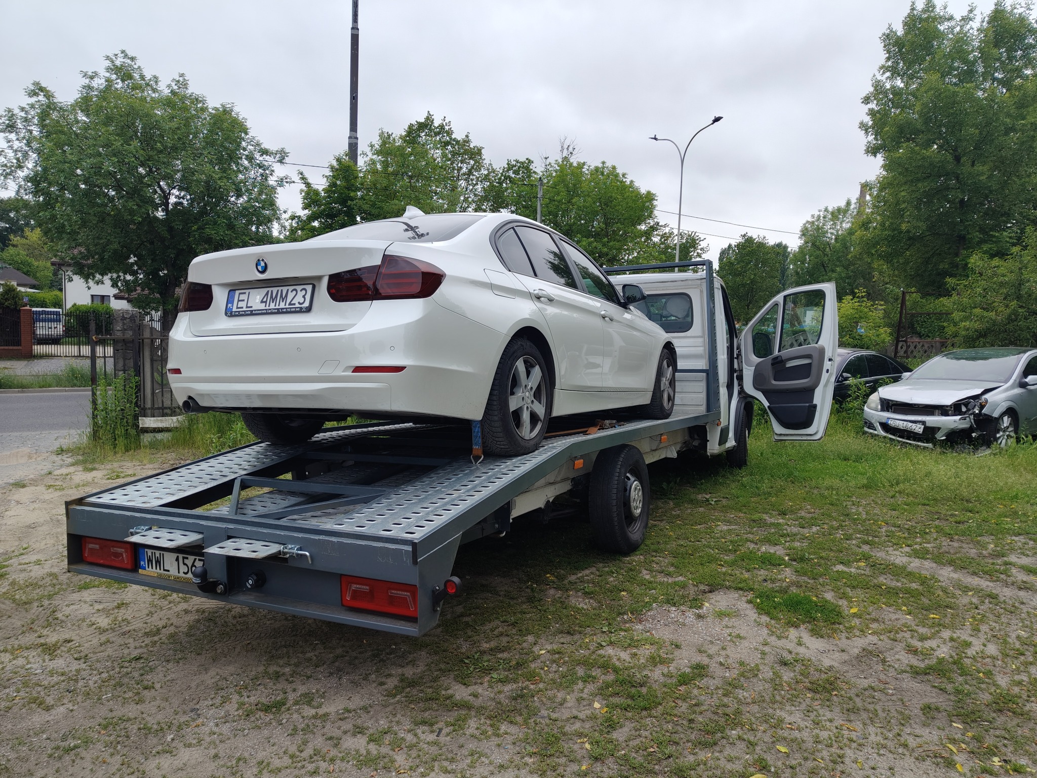 Białe BMW na szarej lawecie z ażurową platformą. W tle uszkodzony pojazd. Otwarta kabina lawety. Tablice rejestracyjne widoczne na obu autach.