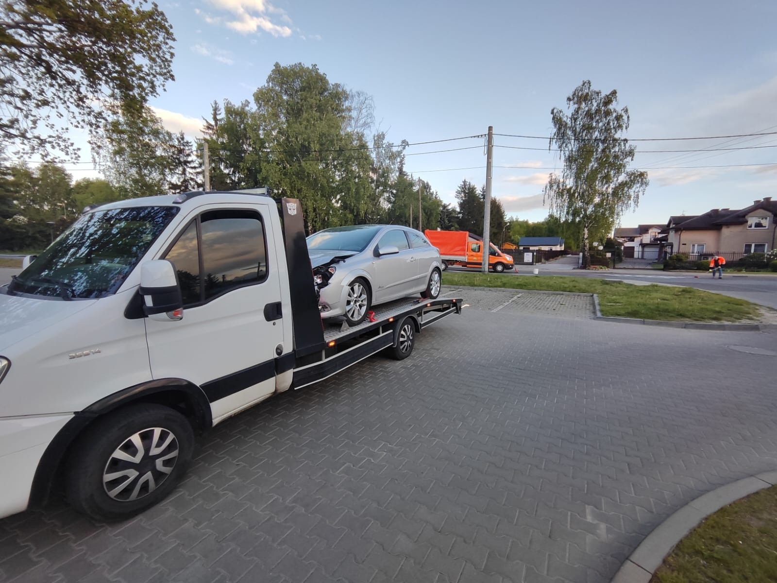 Laweta z uszkodzonym, srebrnym autem na platformie, zaparkowana na brukowanym placu, z pomarańczowym busem w tle i budynkami w oddali. Widok z boku na lawetę.