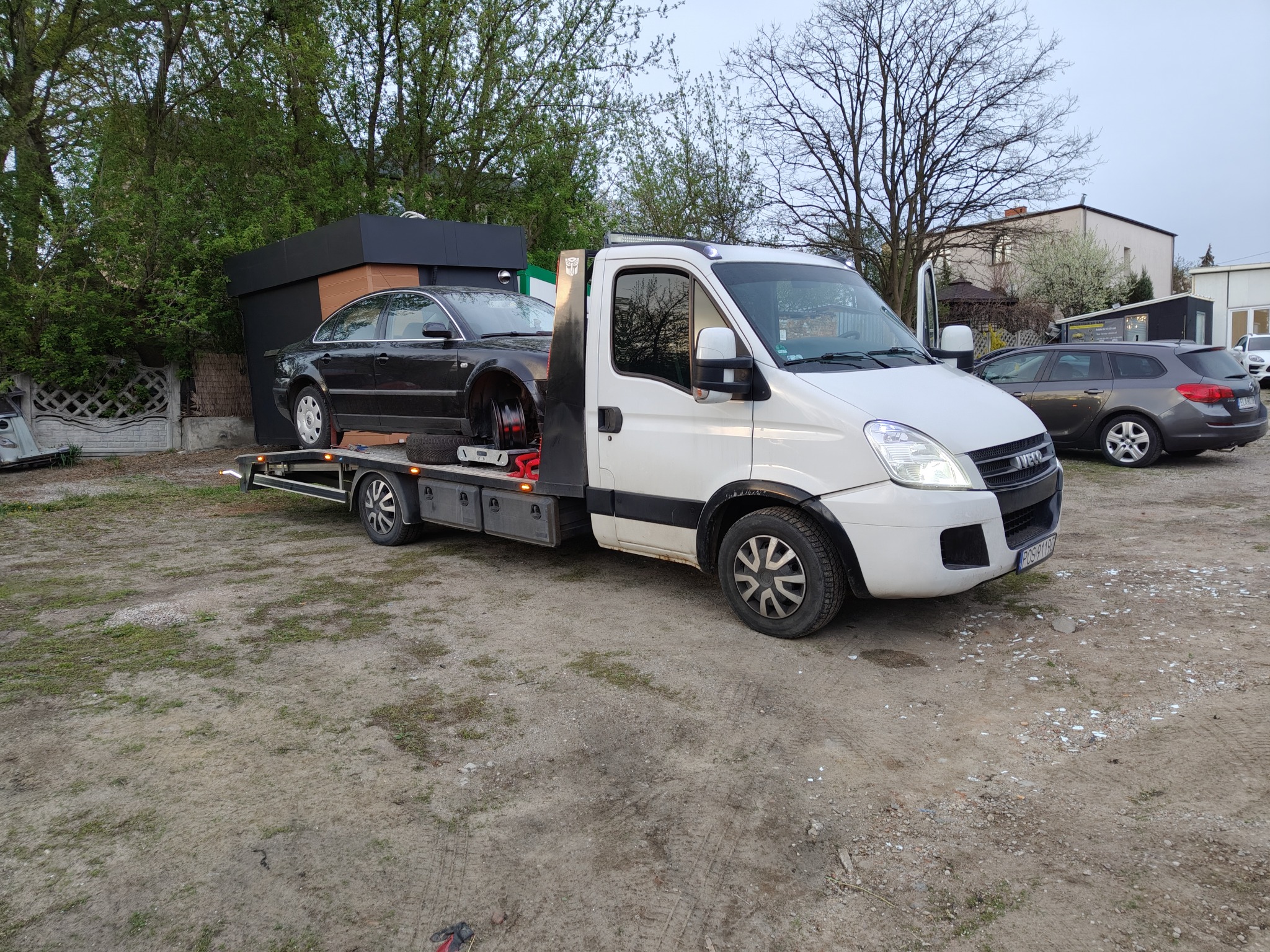 Biała laweta Iveco z czarnym uszkodzonym samochodem osobowym na platformie, zaparkowana na placu. Widoczne budynki w tle i szary samochód osobowy.