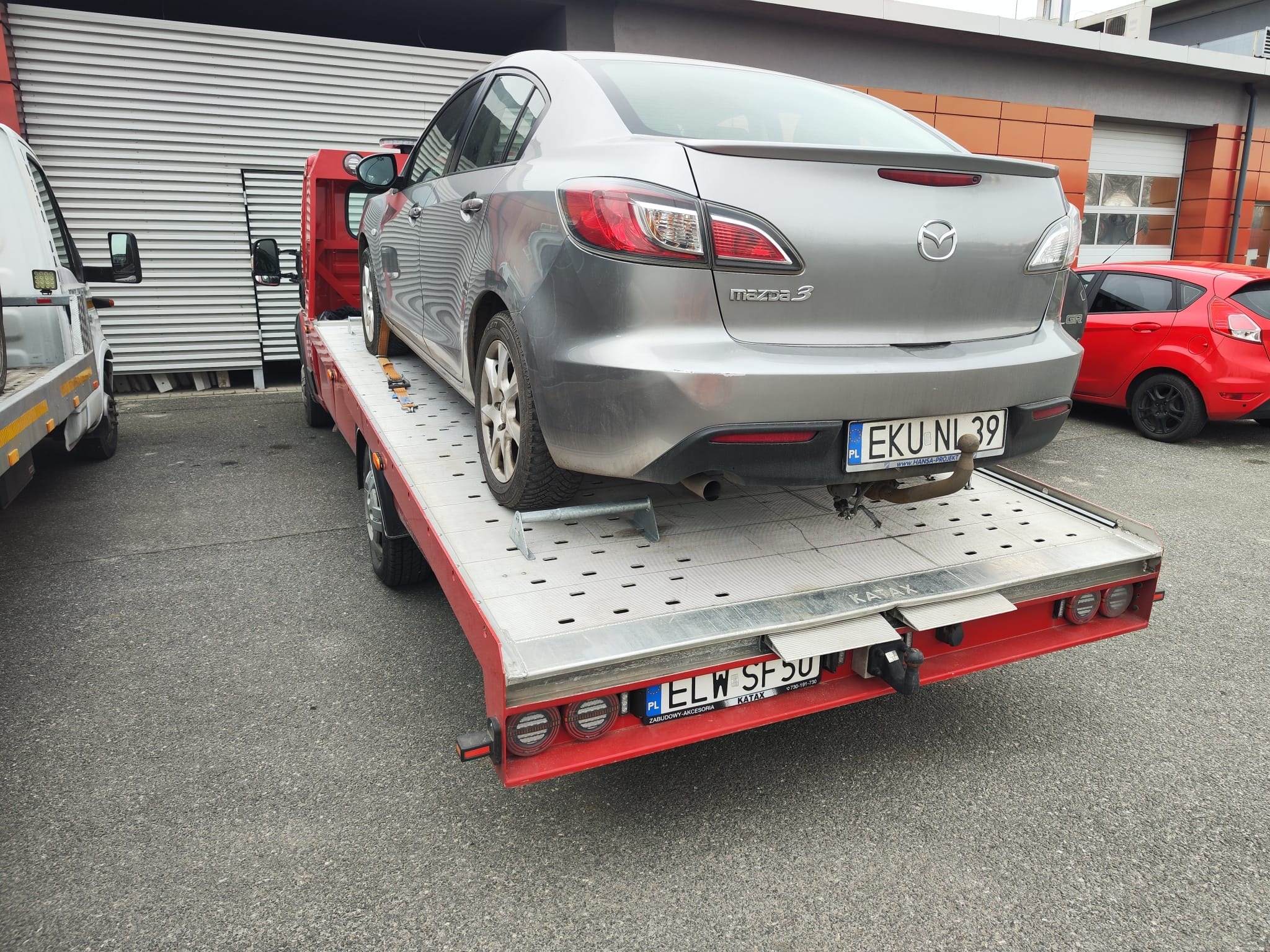 Szara Mazda 3 na czerwonej lawecie z tablicą EKU NI 39, widok z tyłu. W tle budynek z blachy falistej i czerwony samochód. Laweta marki KATAX.