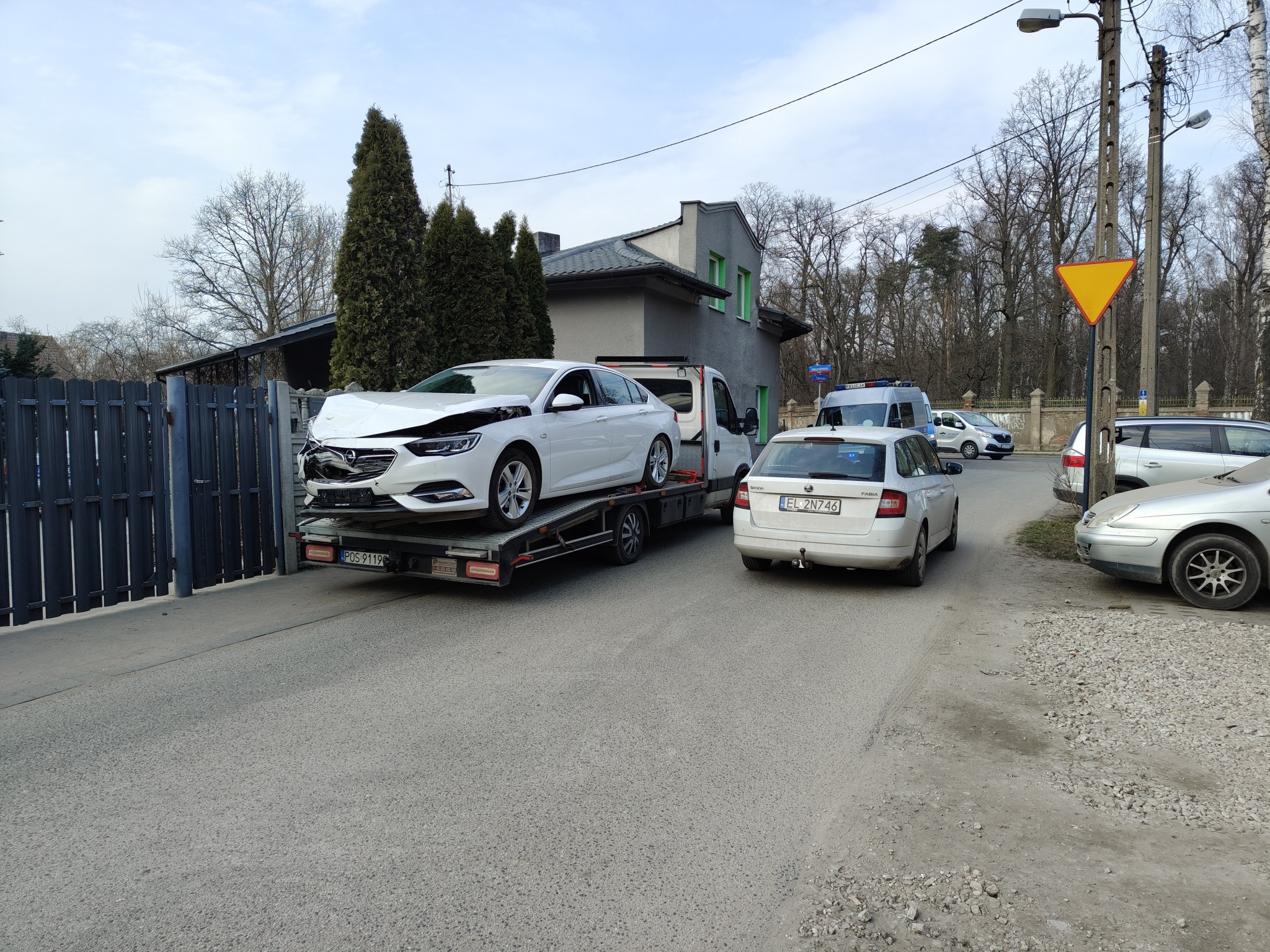 Biała laweta z uszkodzonym białym Oplem Insignia na platformie, zaparkowana na ulicy obok zaparkowanych aut, w tle budynek i drzewa. Widoczny znak drogowy.