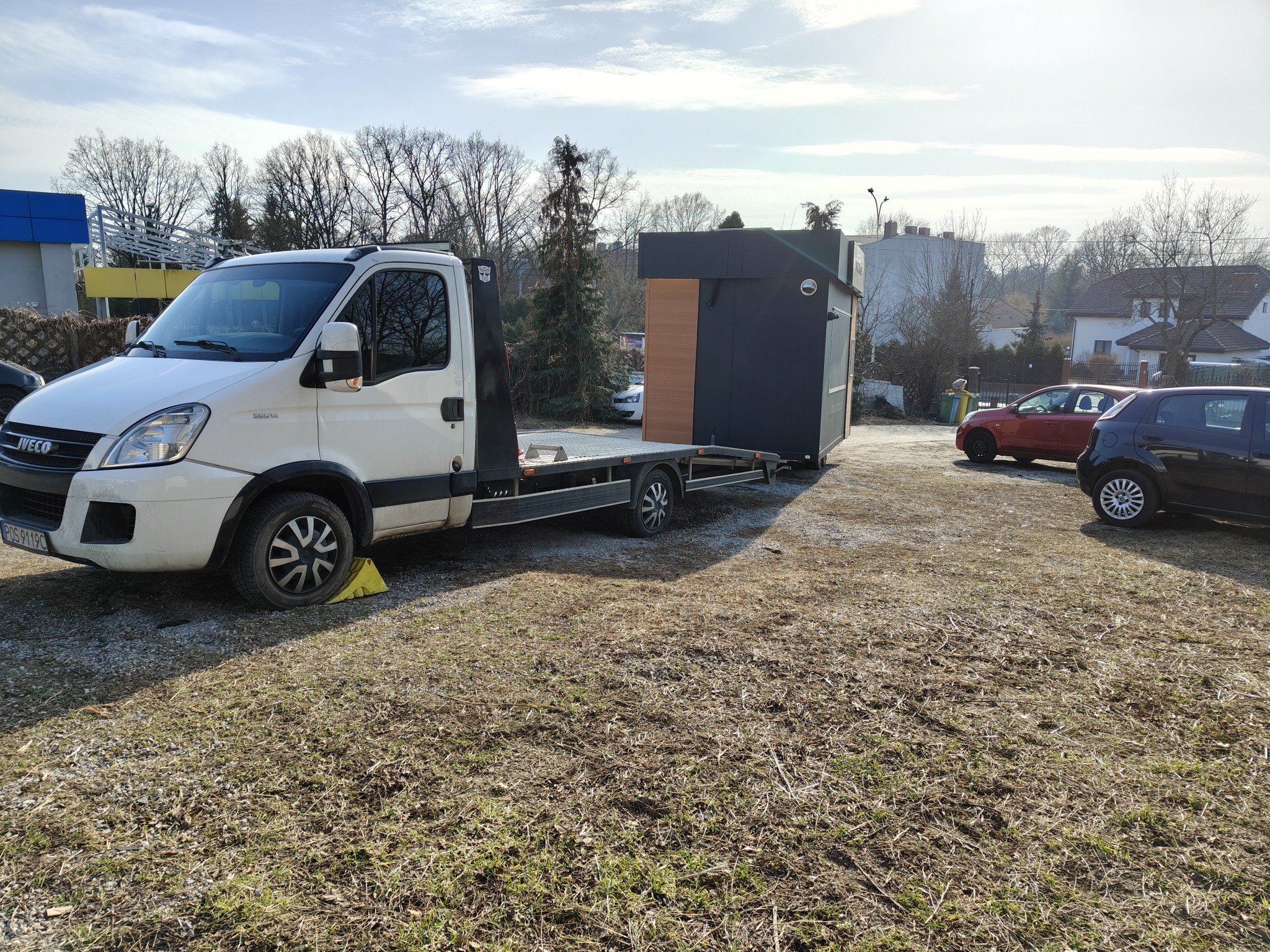 Biała laweta Iveco z żółtym klinem pod kołem przewozi czarny pawilon z elementami drewnianymi, na tle drzew i budynków w słoneczny dzień.