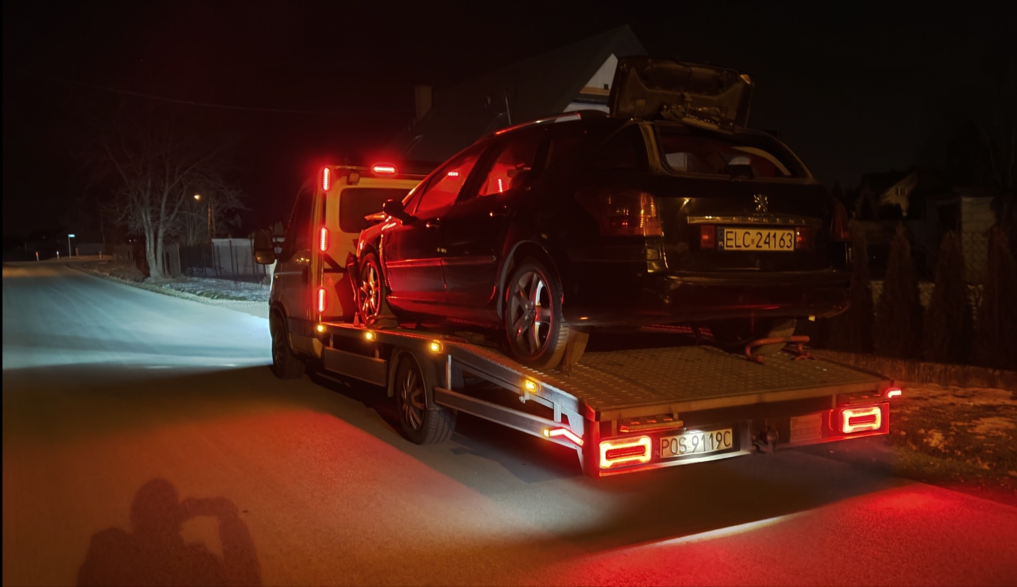 Laweta z czarnym kombi Peugeot na platformie, noc, czerwone światła. Uszkodzony pojazd z otwartą klapą bagażnika, tablica ELC 24163, widok z tyłu.