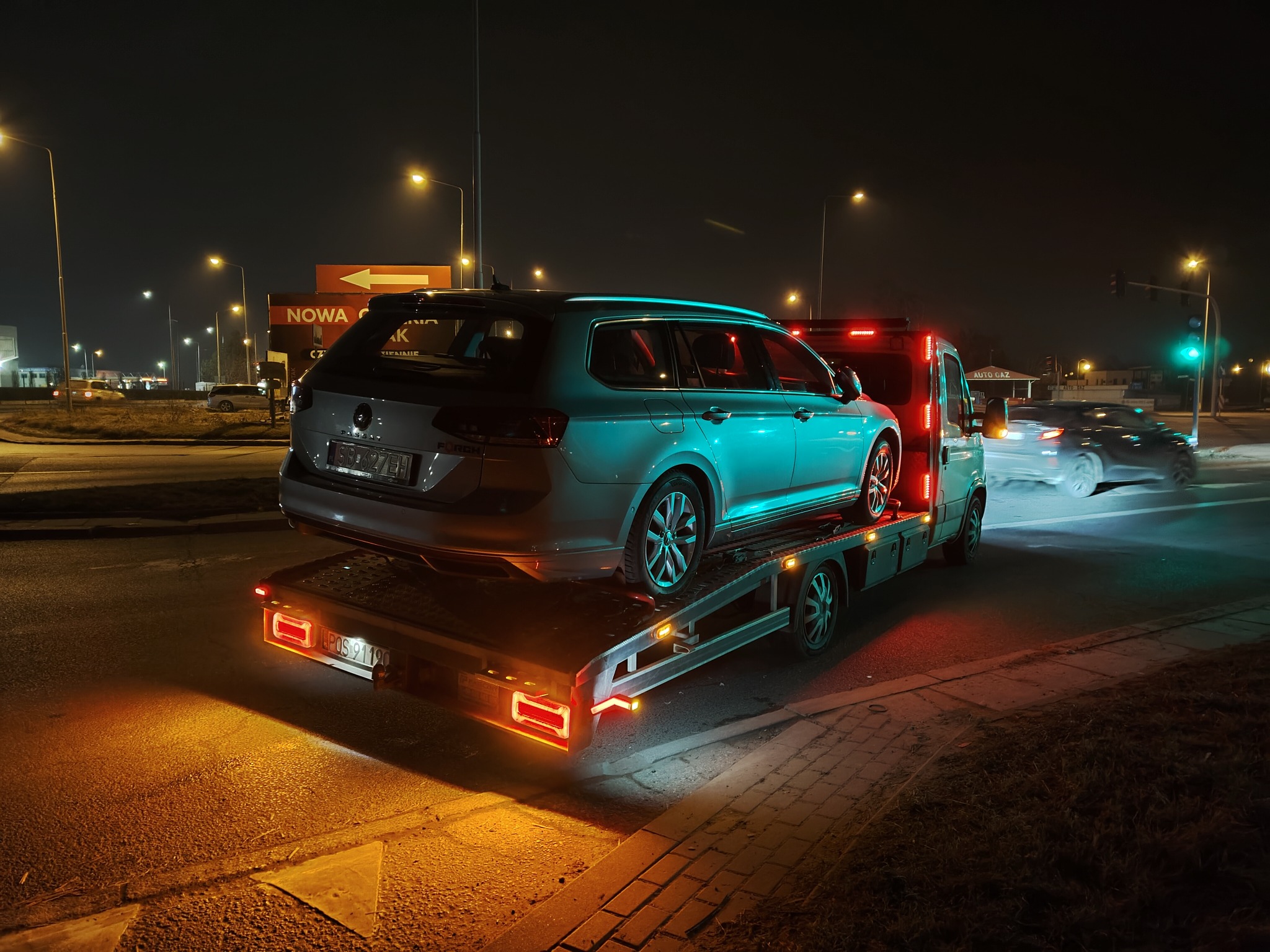 Nocą: szary samochód osobowy na platformie lawety z włączonym oświetleniem ostrzegawczym. W tle ulica miejska z latarniami i budynkami.