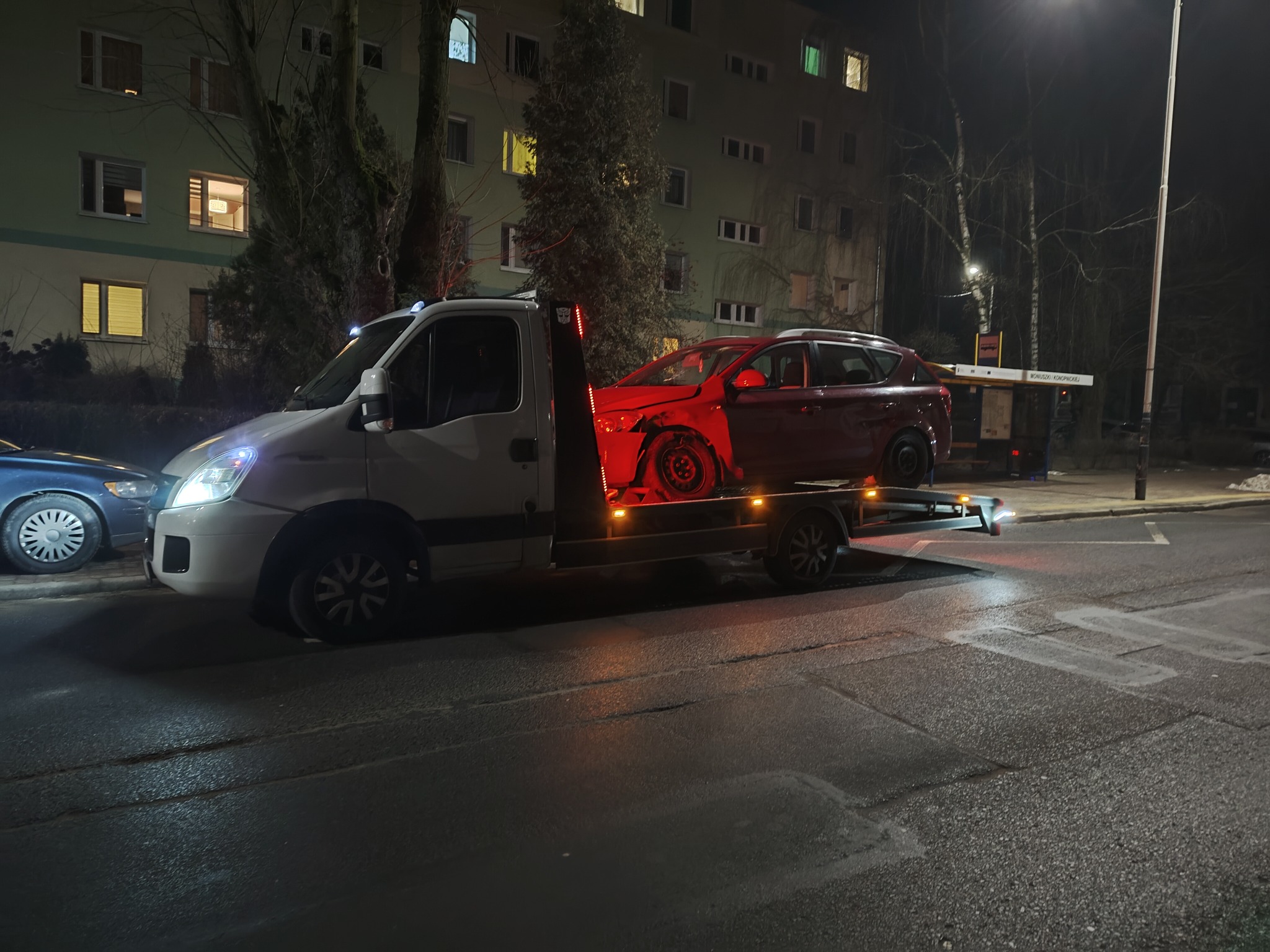 Nocna scena z lawetą przewożącą uszkodzony, czerwony samochód osobowy na tle bloku mieszkalnego. Oświetlenie uliczne. Widoczny fragment zaparkowanego auta.