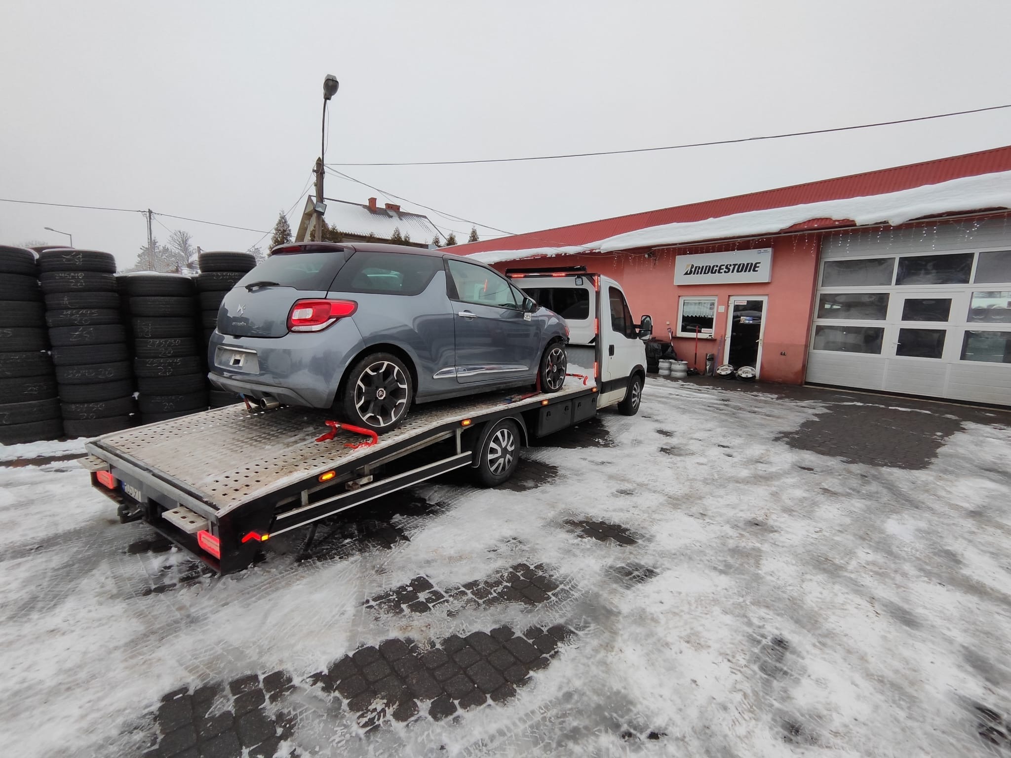 Szary Citroen DS3 na platformie lawety, z tyłu budynek serwisu Bridgestone. Zimowa sceneria z oponami w tle. Ujęcie z boku.
