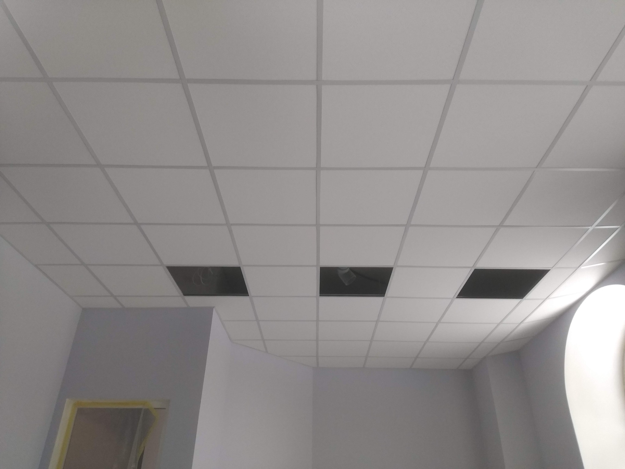 Biały sufit kasetonowy z widocznymi otworami na instalacje, szare ściany w pomieszczeniu. Widok z dołu, wnętrze w trakcie prac wykończeniowych.