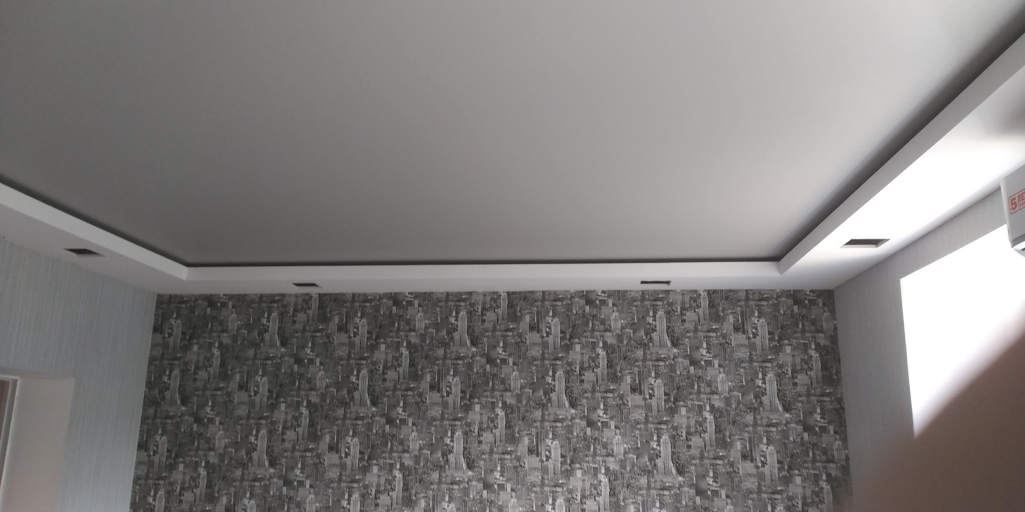 Wykończony sufit podwieszany z oświetleniem punktowym i ściana z tapetą w motywie miejskim. Widok na róg pokoju, z fragmentem klimatyzacji po prawej.