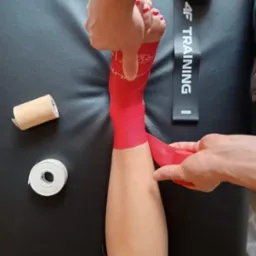 Nakładanie czerwonej taśmy kinesiotaping na stopę pacjenta na czarnym tle, widoczne rolki taśmy i piłka do masażu. Kąt z góry.