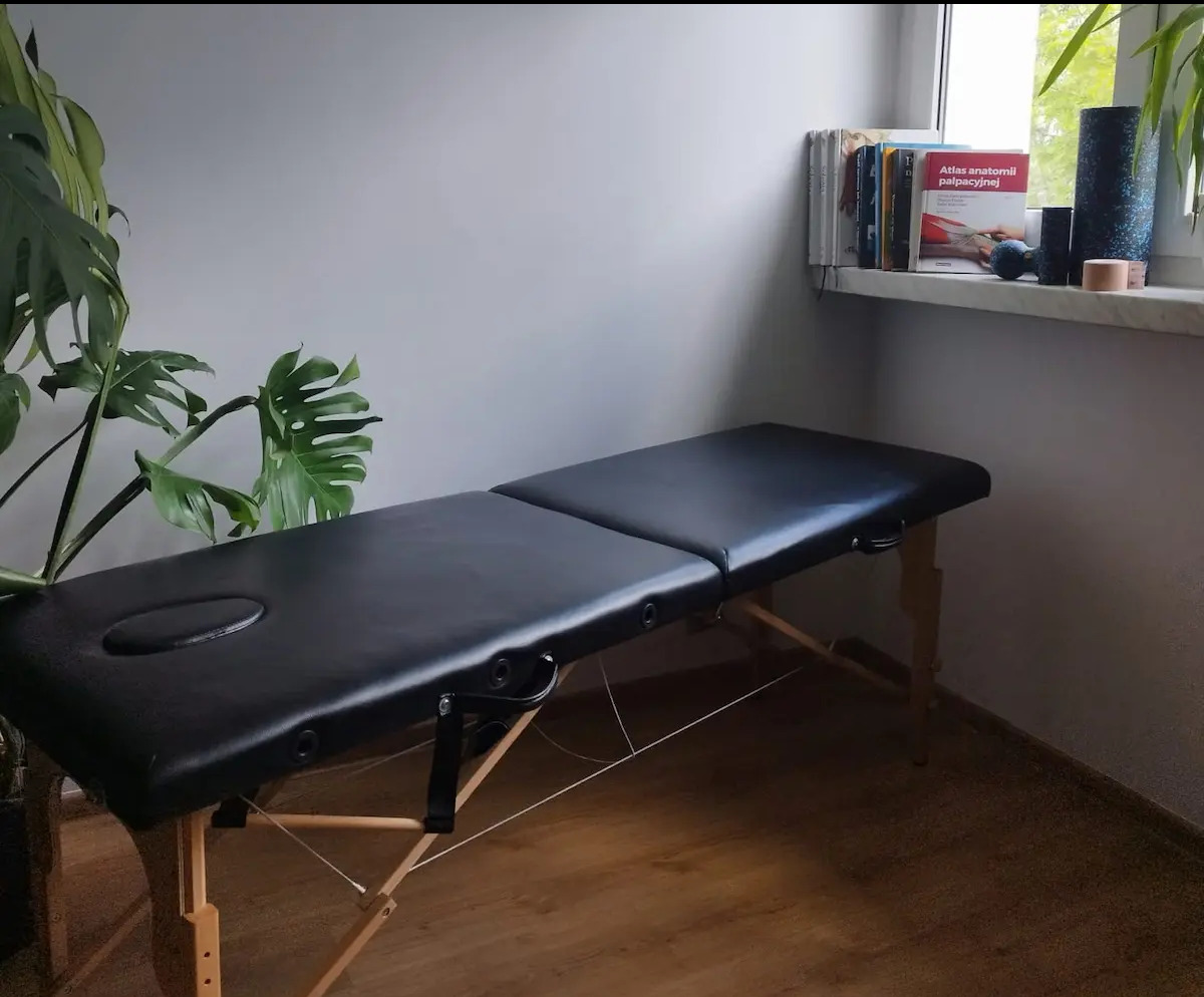 Profesjonalne łóżko do masażu z czarną tapicerką, ustawione w gabinecie z elementami roślinnymi i książkami anatomicznymi na parapecie okiennym.