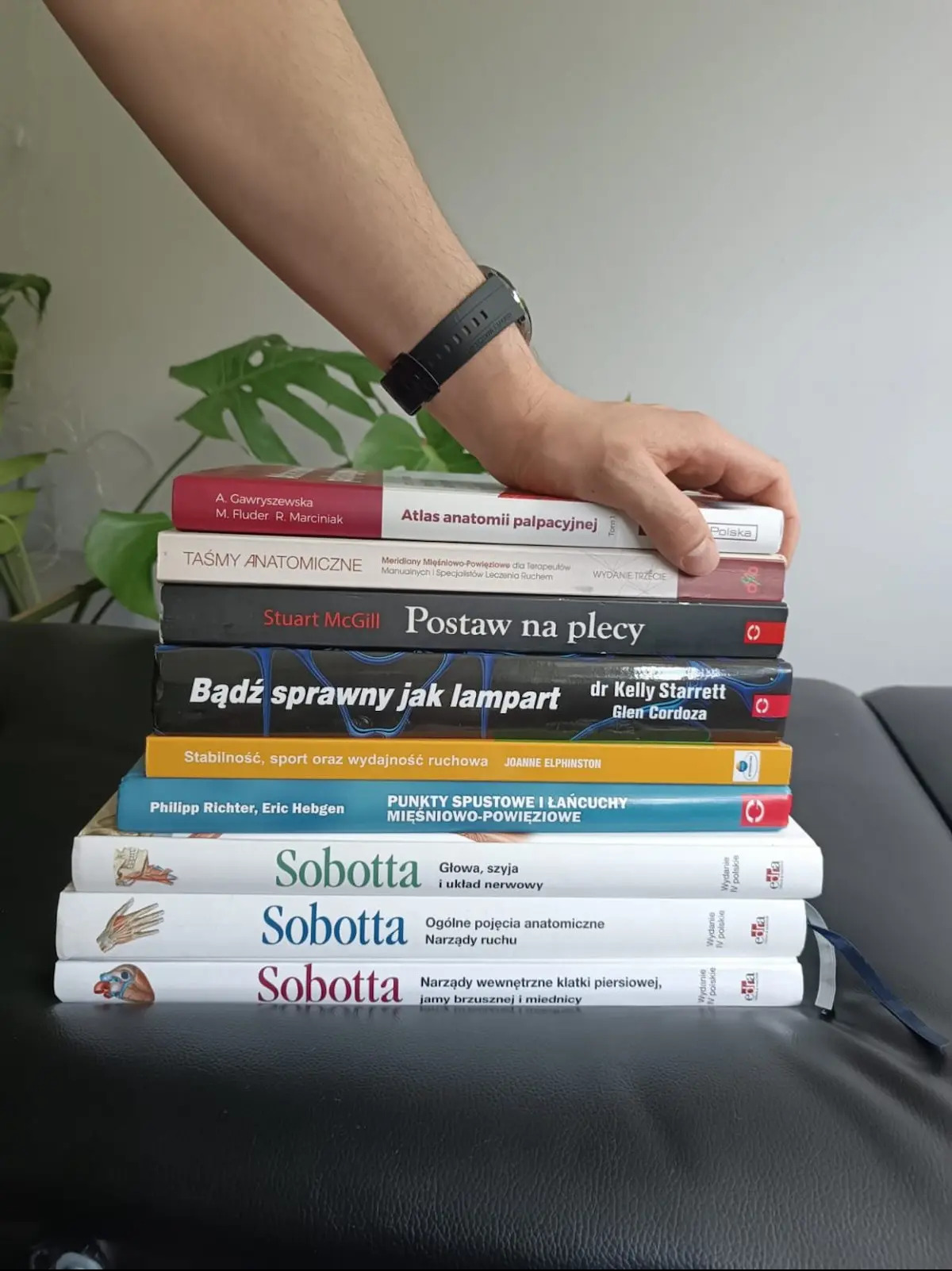 Stos książek o anatomii i fizjoterapii, z dłonią na wierzchu, na czarnej powierzchni. Widoczne tytuły: Atlas anatomii palpacyjnej, Taśmy anatomiczne, Postaw na plecy.