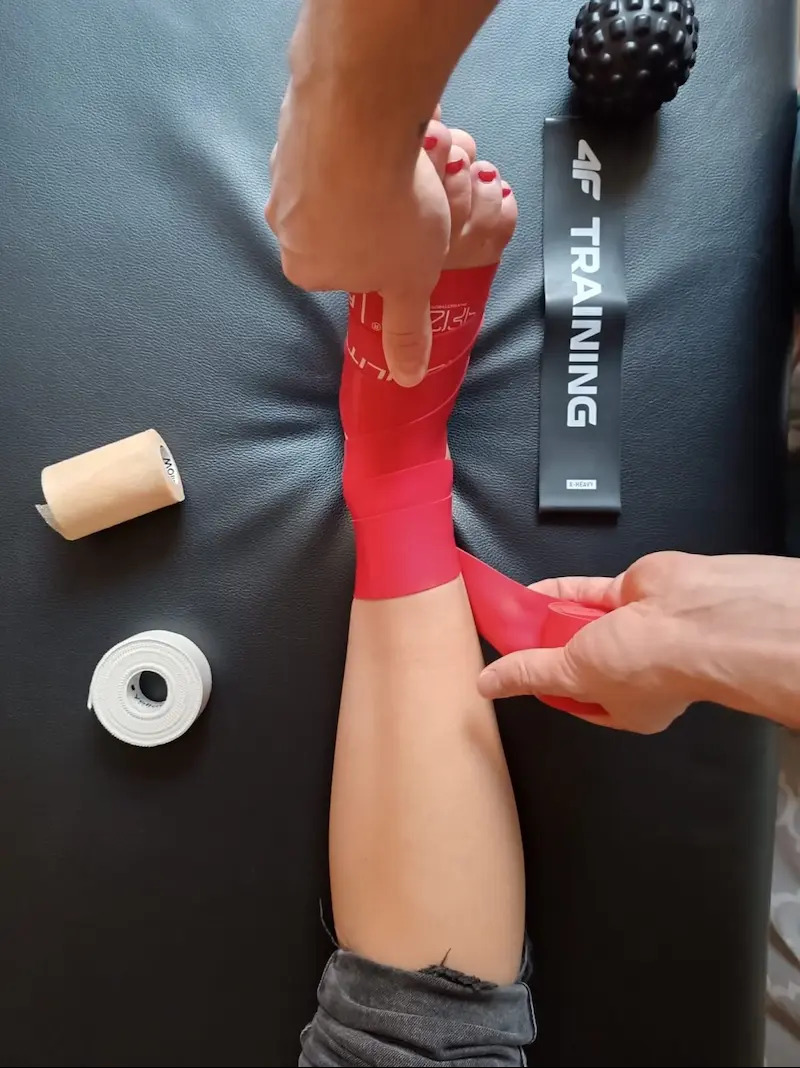 Nakładanie czerwonej taśmy kinesiotaping na stopę pacjenta na czarnym tle, widoczne rolki taśmy i piłka do masażu. Kąt z góry.