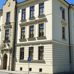 Elewacja budynku biurowego z jasną fasadą i ciemnymi ramami okiennymi w słoneczny dzień. Widoczny znak drogowy i fragment zaparkowanego samochodu.