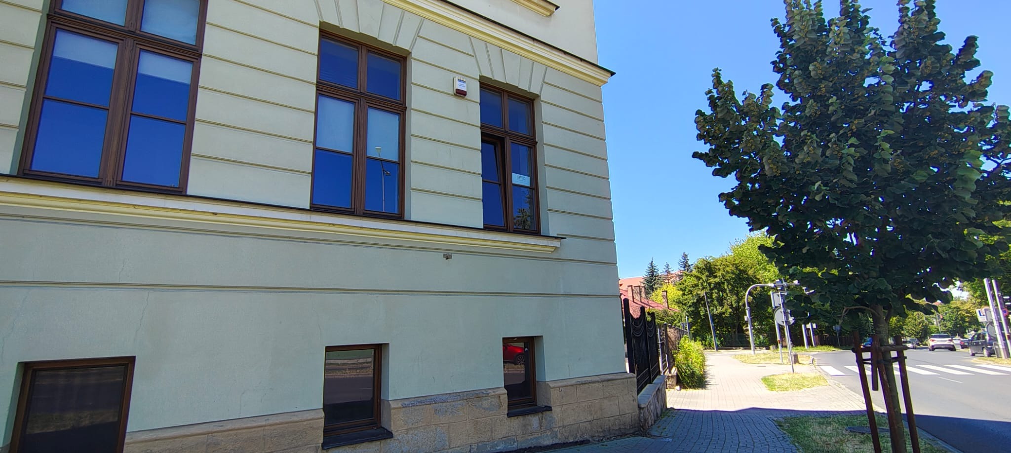 Elewacja budynku biurowego z oknami w brązowych ramach, widok z zewnątrz, błękitne niebo. Na jednym z okien naklejka informacyjna.