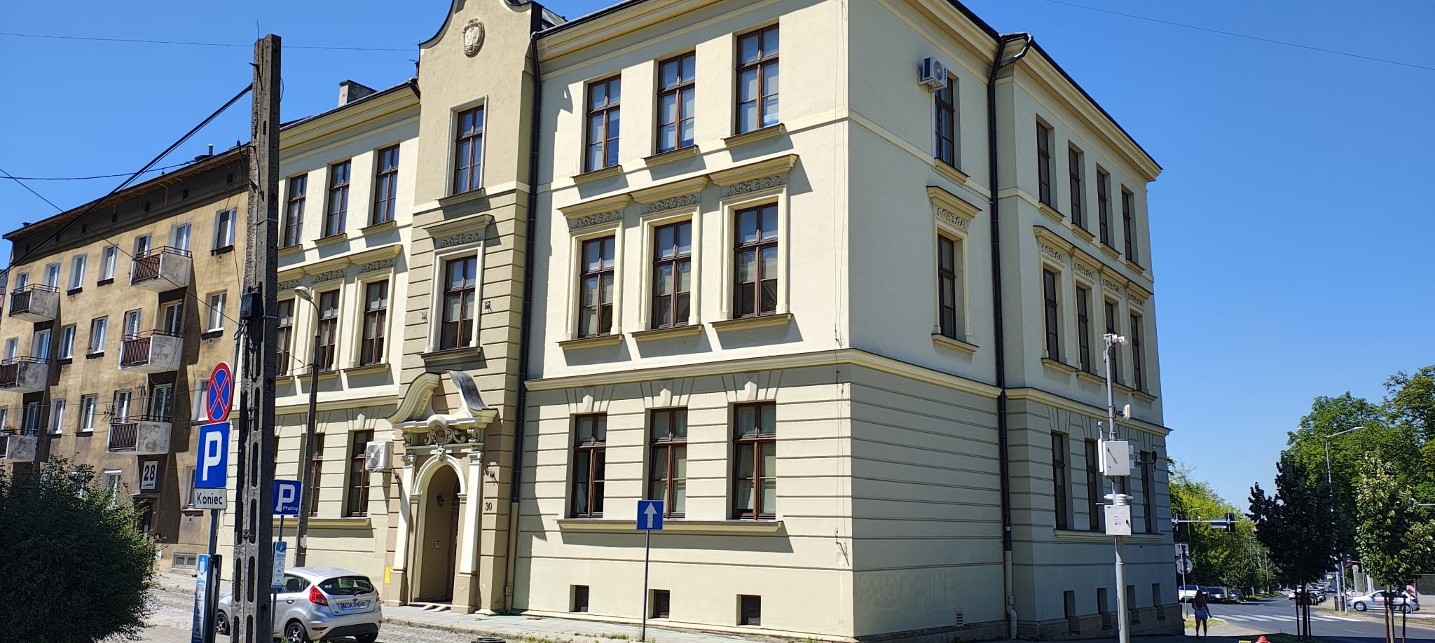 Elewacja budynku biurowego z jasną fasadą i ciemnymi ramami okiennymi w słoneczny dzień. Widoczny znak drogowy i fragment zaparkowanego samochodu.