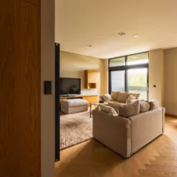 Jasne, przestronne wnętrze salonu z parkietem w jodełkę, beżową sofą i dużym oknem balkonowym. Widok przez otwarte drzwi na telewizor i meble.