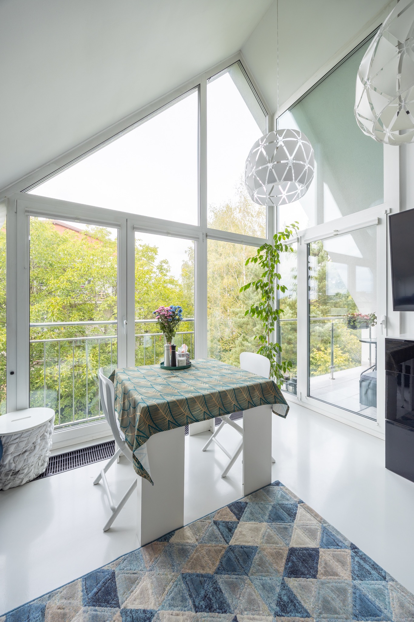 Jasna jadalnia z dużymi oknami i balkonem. Stół z wzorzystym obrusem, kwiaty, dywan w geometryczne wzory, nowoczesne lampy. Widok na zieleń za oknem.